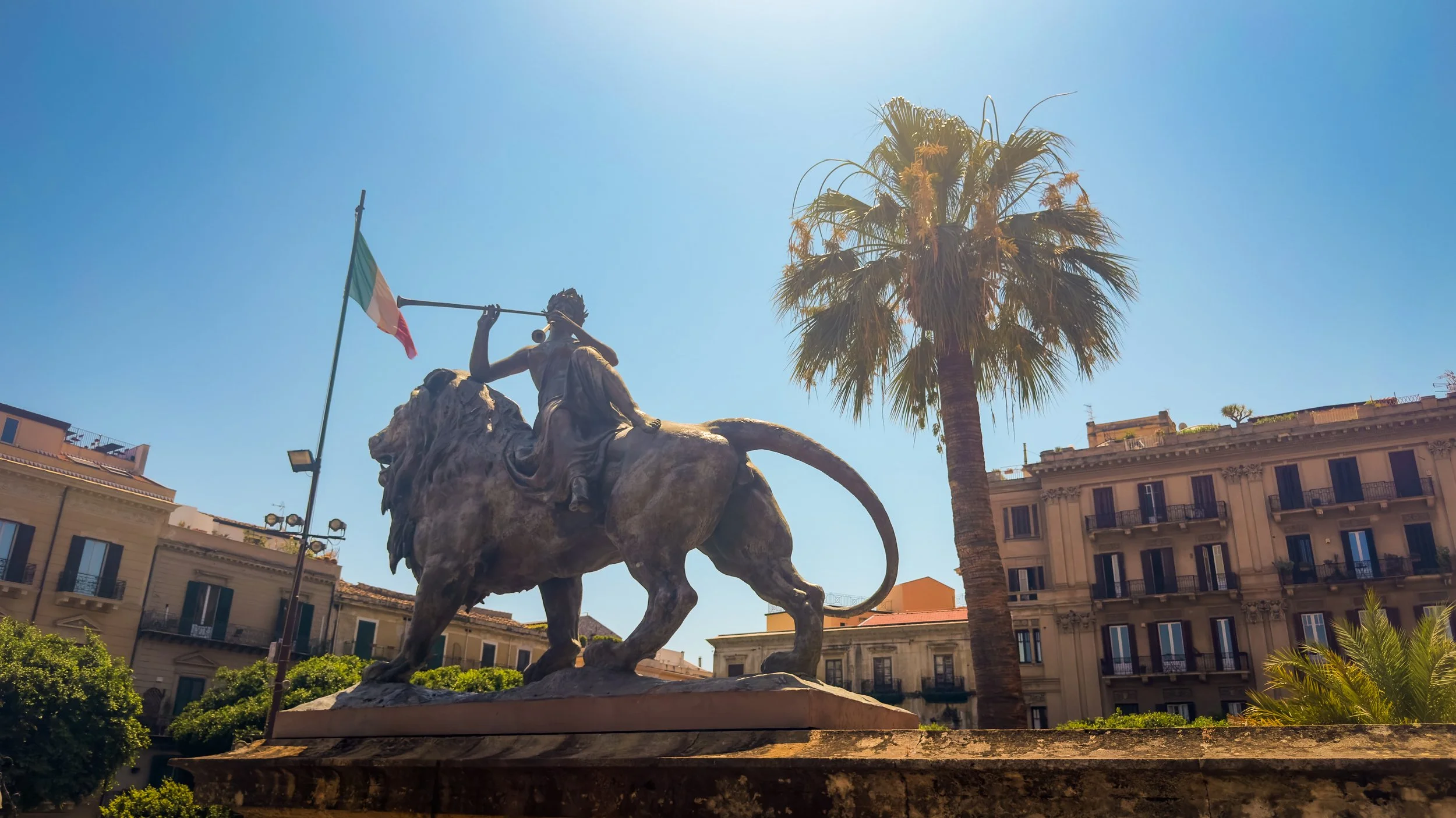 Bronze lion topped by the allegorical figure of La Lirica outside Teatro Massimo in Palermo, with a palm tree and Italian flag behind.