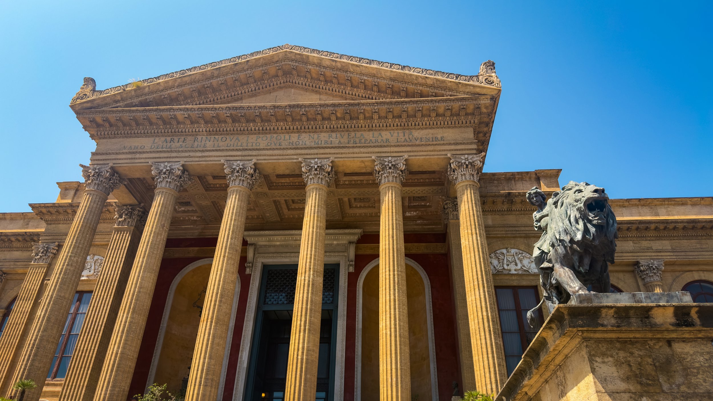 Exterior of Teatro Massimo in Palermo with tall columns and a lion sculpture beneath a clear blue sky.