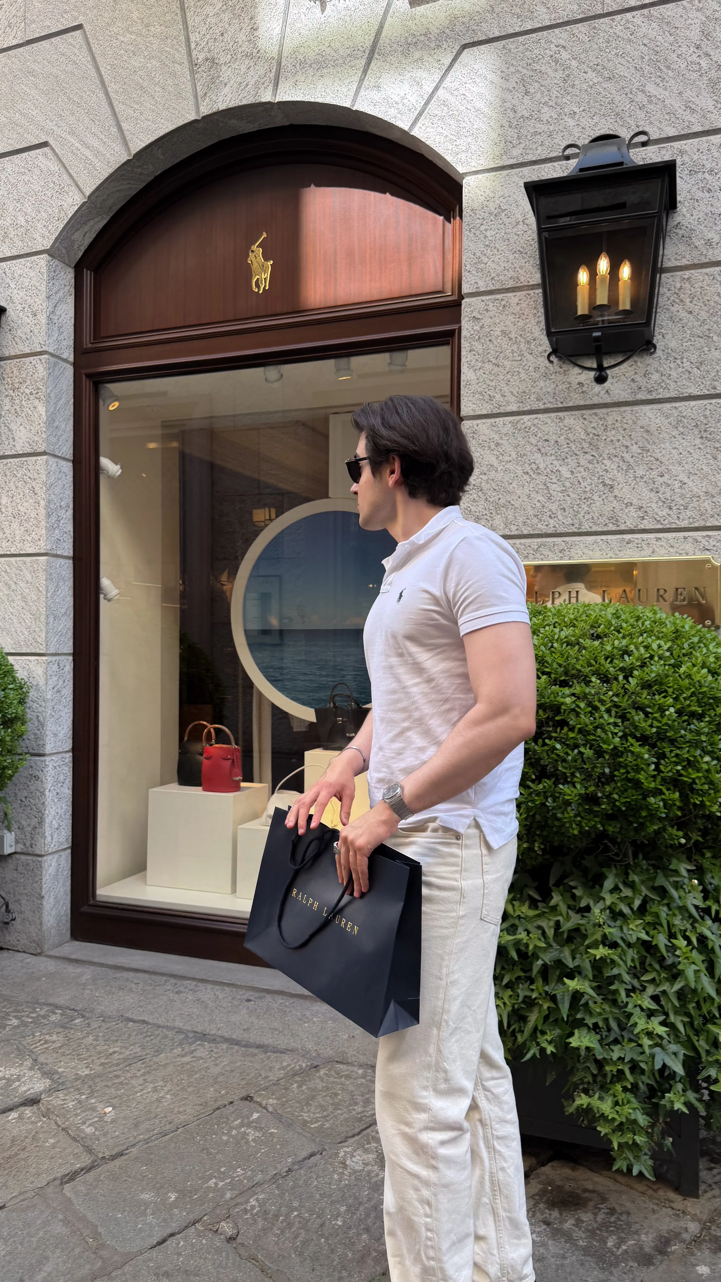 A man wearing sunglasses, a white Polo Ralph Lauren shirt, and beige pants holding a Ralph Lauren shopping bag standing outside a storefront with a display window featuring handbags and a background of a city sidewalk.