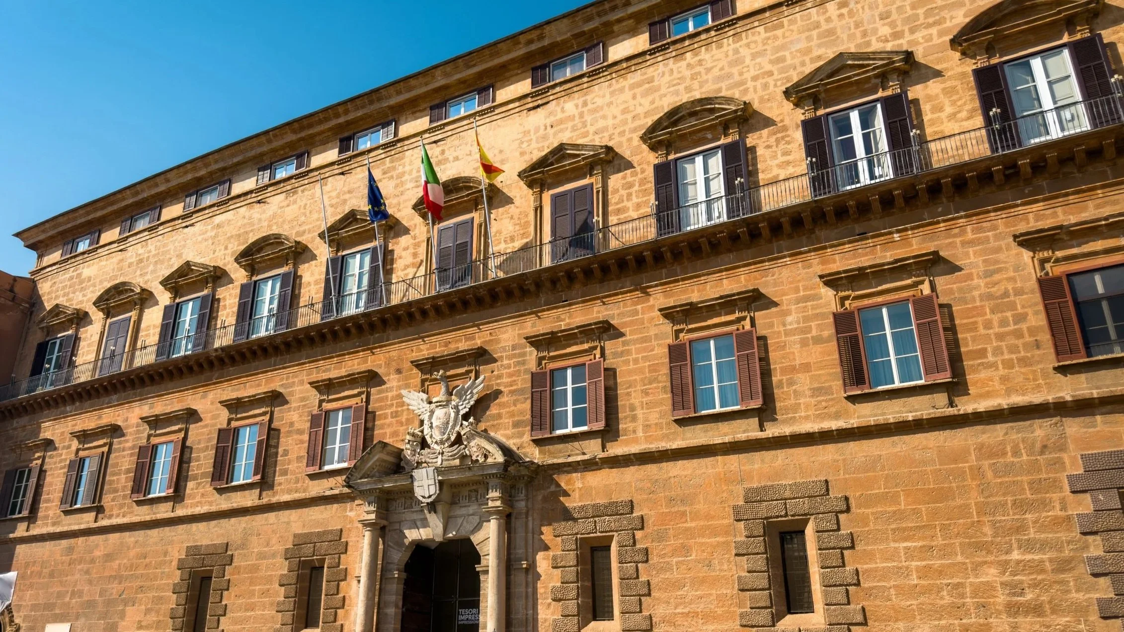 Stone façade of Palazzo dei Normanni in Palermo with shuttered windows and flags above the main entrance.