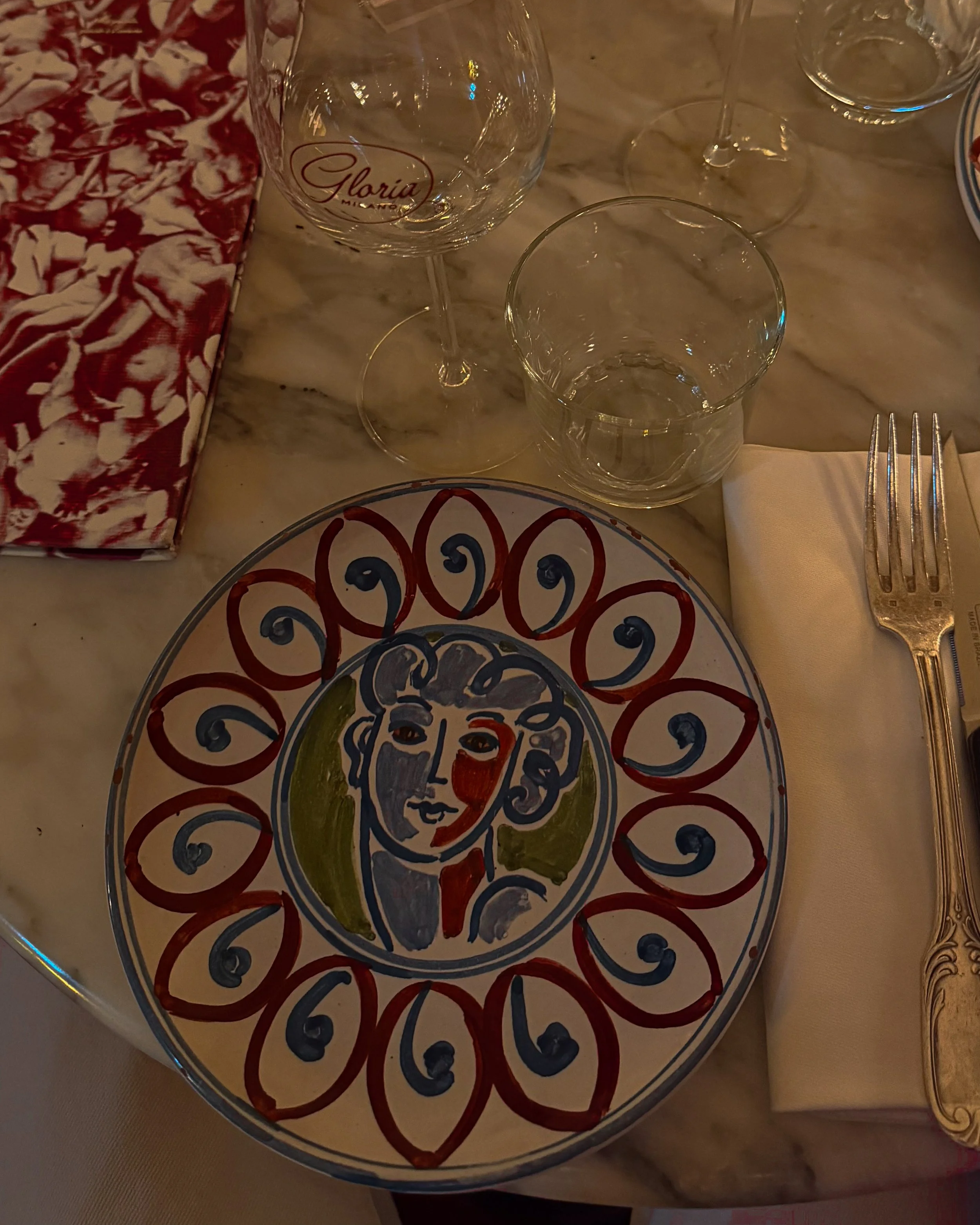 Table setting at Gloria Osteria in Milan with decorative plate, branded wine glass, marble tabletop, and red patterned napkin.
