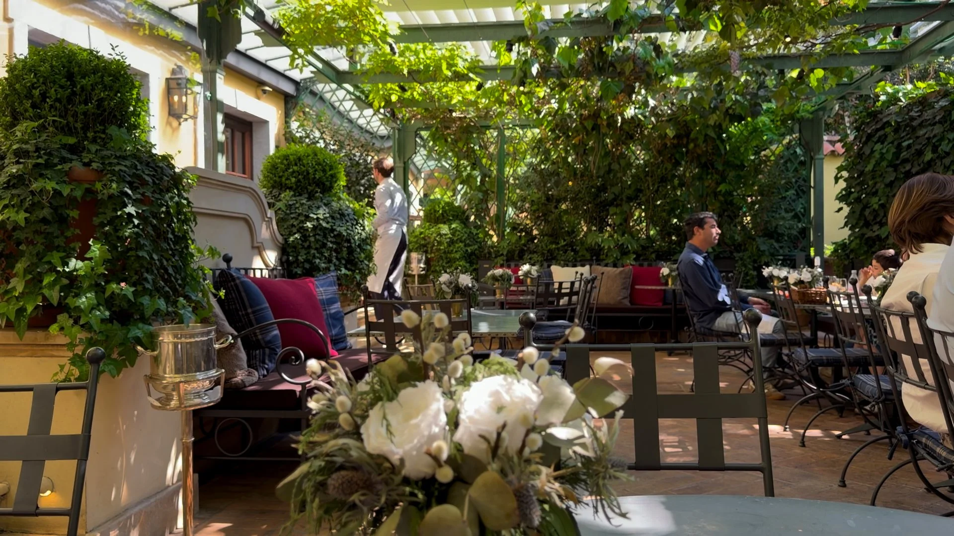 Leafy outdoor garden dining area at Ralph’s Bar Milano with iron furniture, flowers and guests seated beneath a vine-covered pergola.