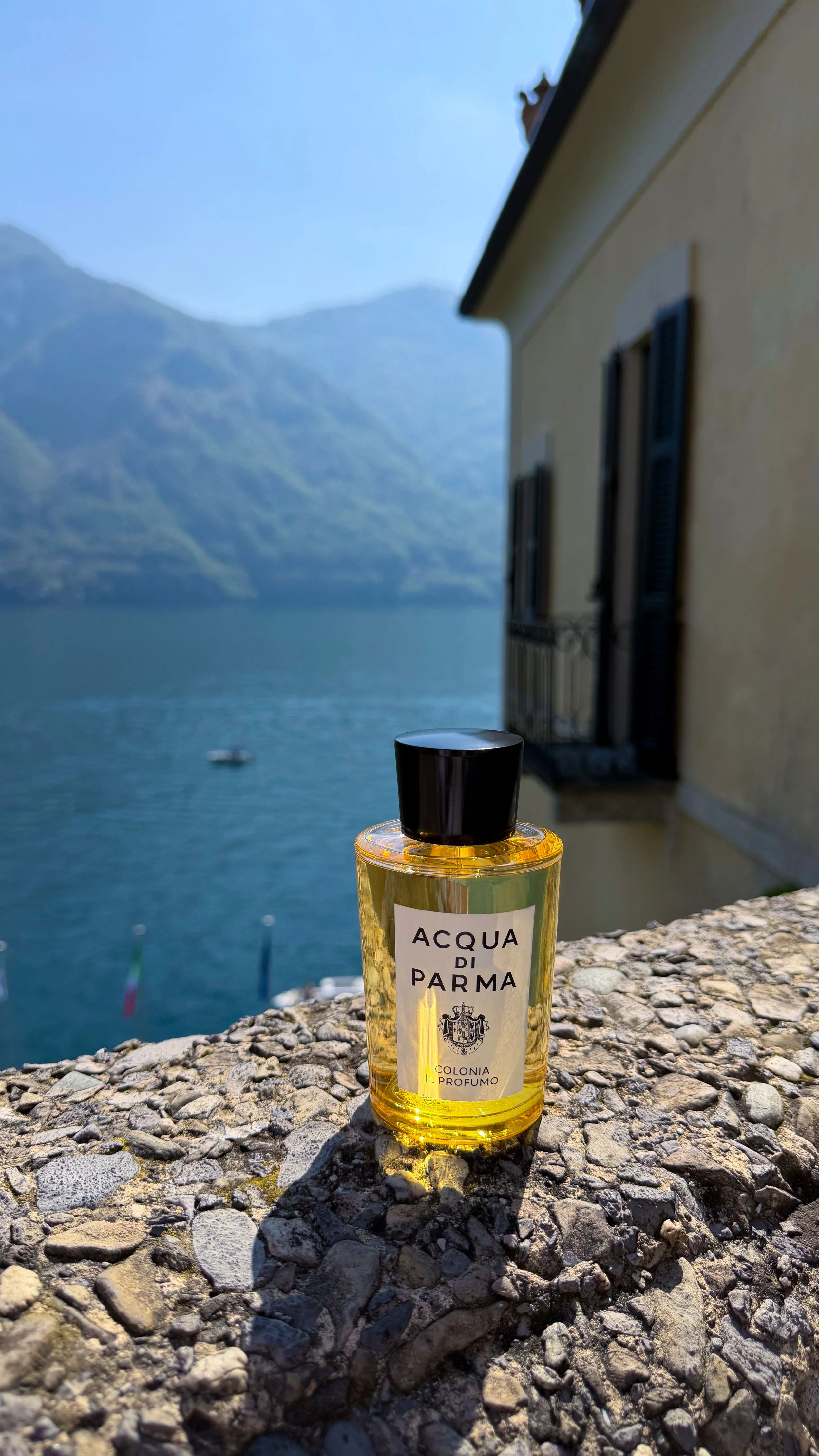 A bottle of Acqua di Parma perfume placed on a stone surface with a scenic view of water and mountains in the background.