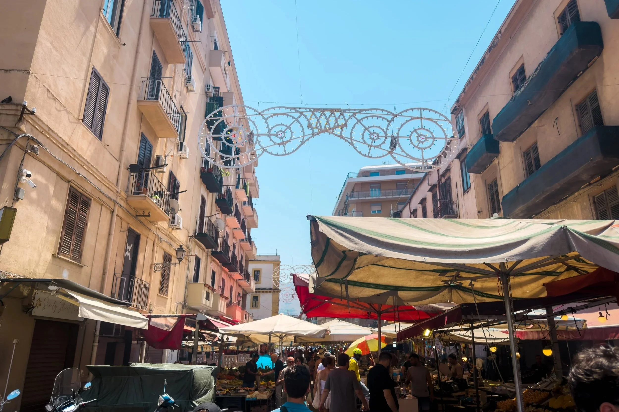 Busy street at Mercato Ballarò in Palermo with produce stalls, canvas awnings and pedestrians.
