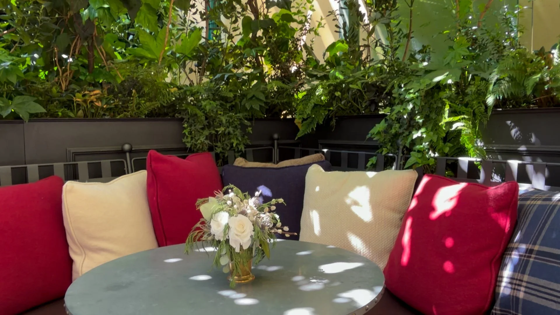 Garden banquette at Ralph’s Bar Milano with red, cream and navy cushions, a round table, flowers and dense greenery.
