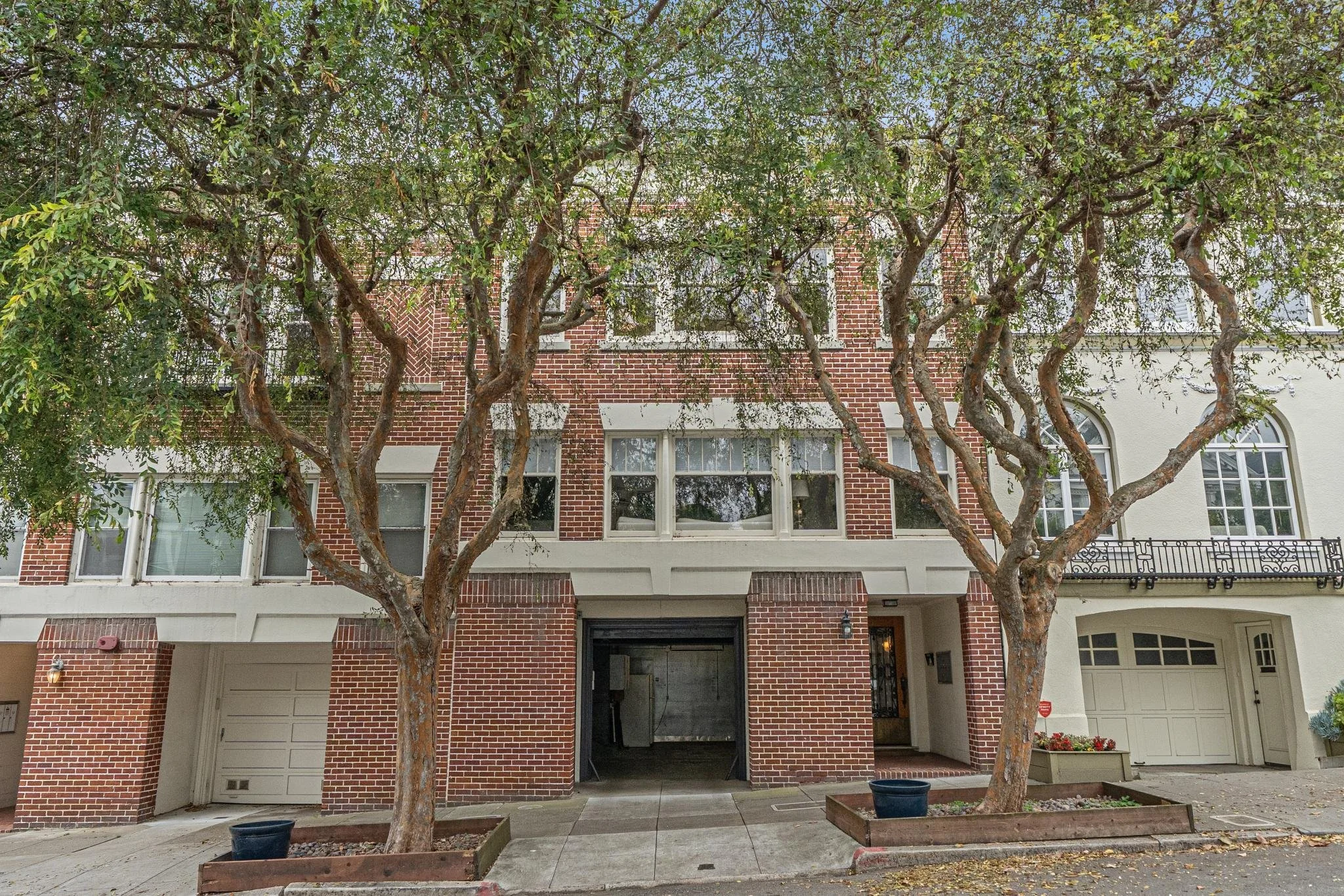 A brick apartment building with multiple windows, two trees with twisted branches in front, and a sidewalk and driveway area.