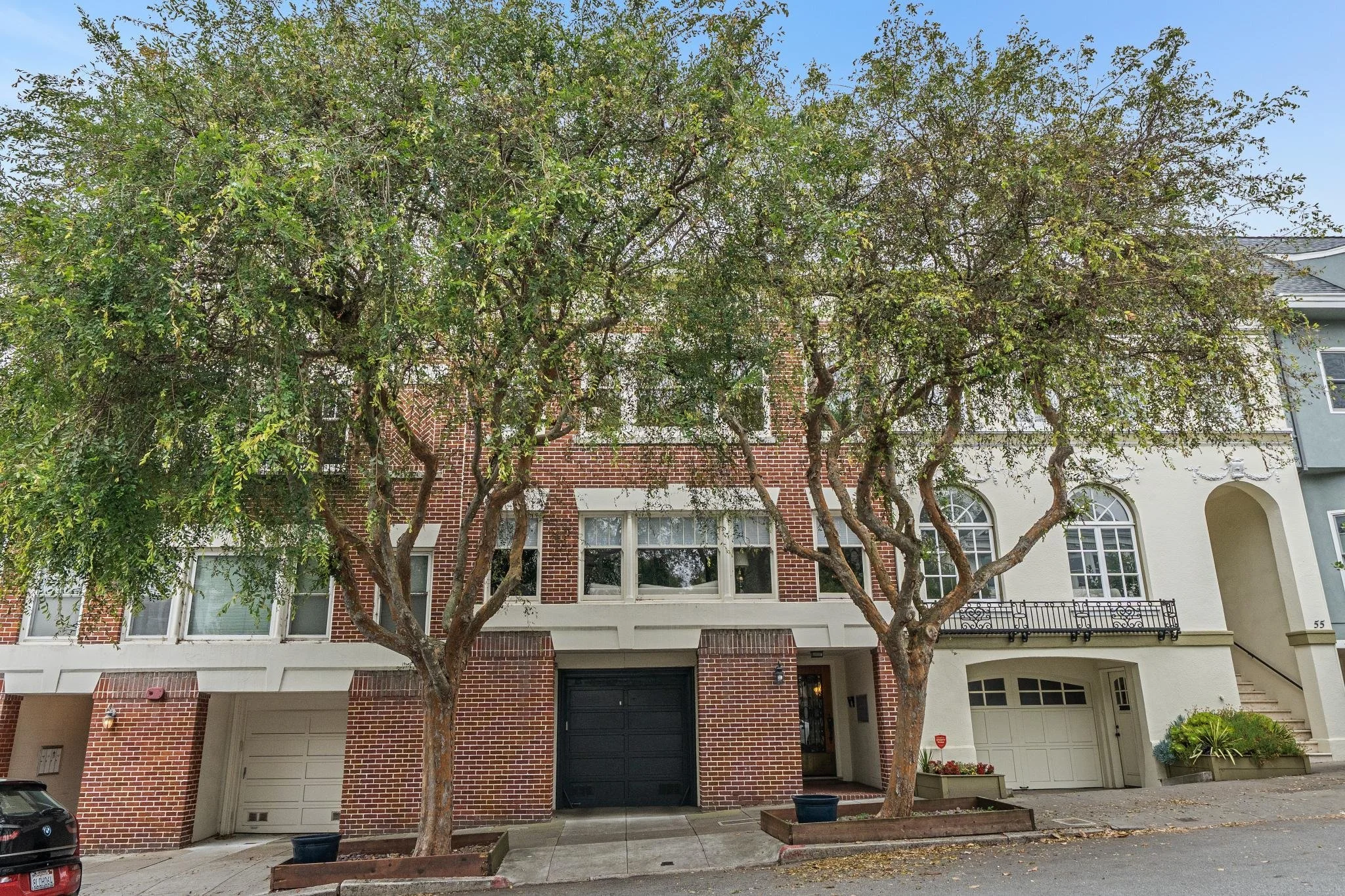 Two large trees with green foliage are in front of a multi-story brick and light-colored residential building. The building has several large windows, and there are two garages at the ground level. The driveway and sidewalk are visible, with some pot