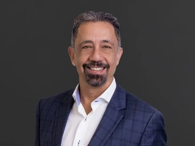 A middle-aged man with short dark hair, a goatee, and a mustache, smiling in a professional portrait against a gray background, wearing a navy blue checkered blazer and a white shirt.