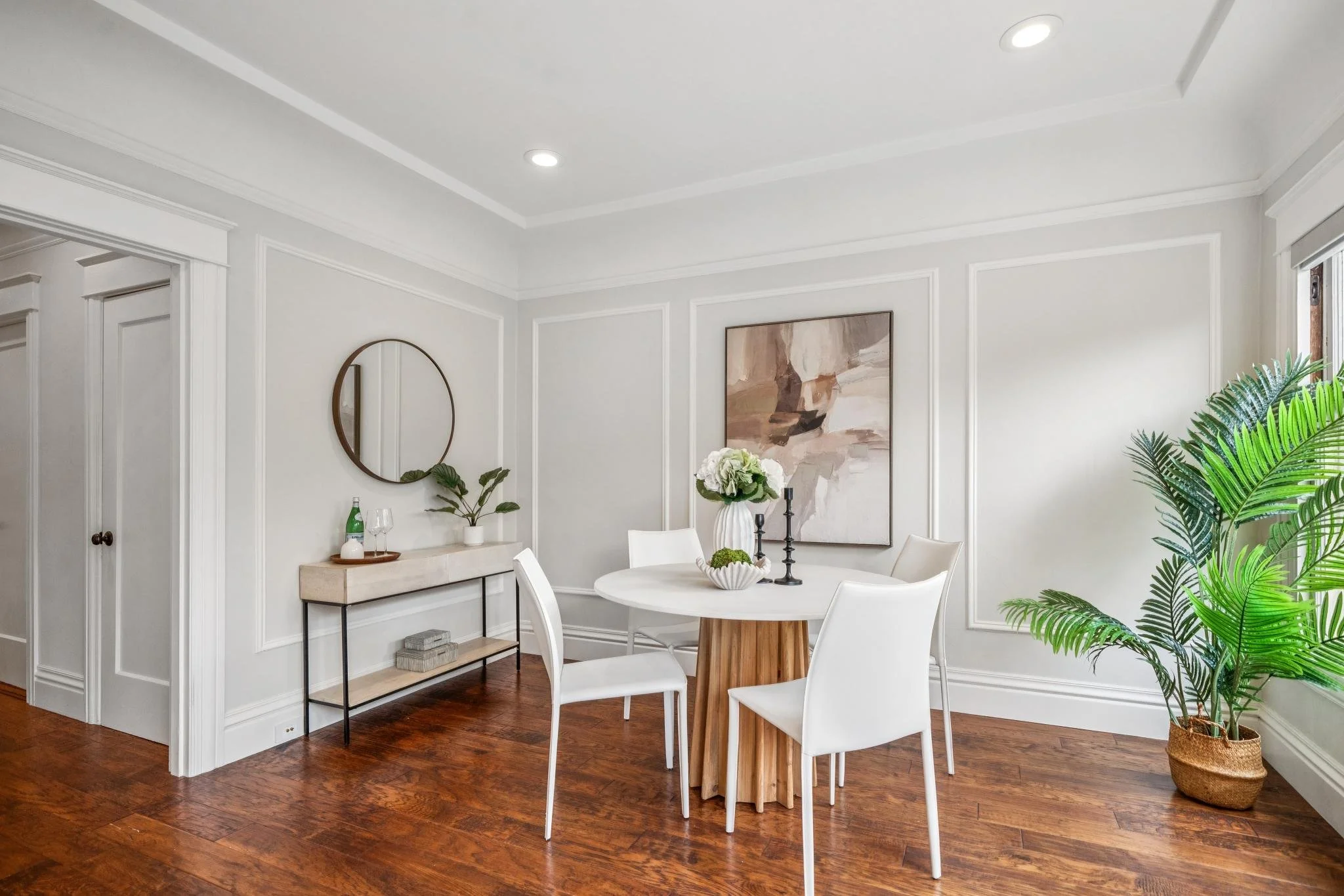 A dining area with white walls and wood flooring, featuring a round white table with white chairs, a side table with a potted plant and decorative items, a large abstract painting, and tall green plants near a window.