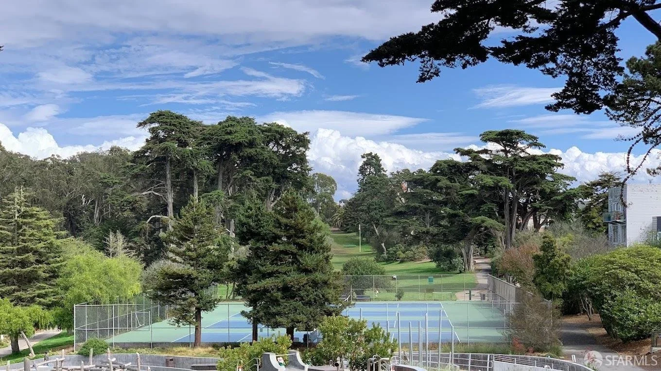 View of a park with tennis courts, large green trees, and a partly cloudy sky.