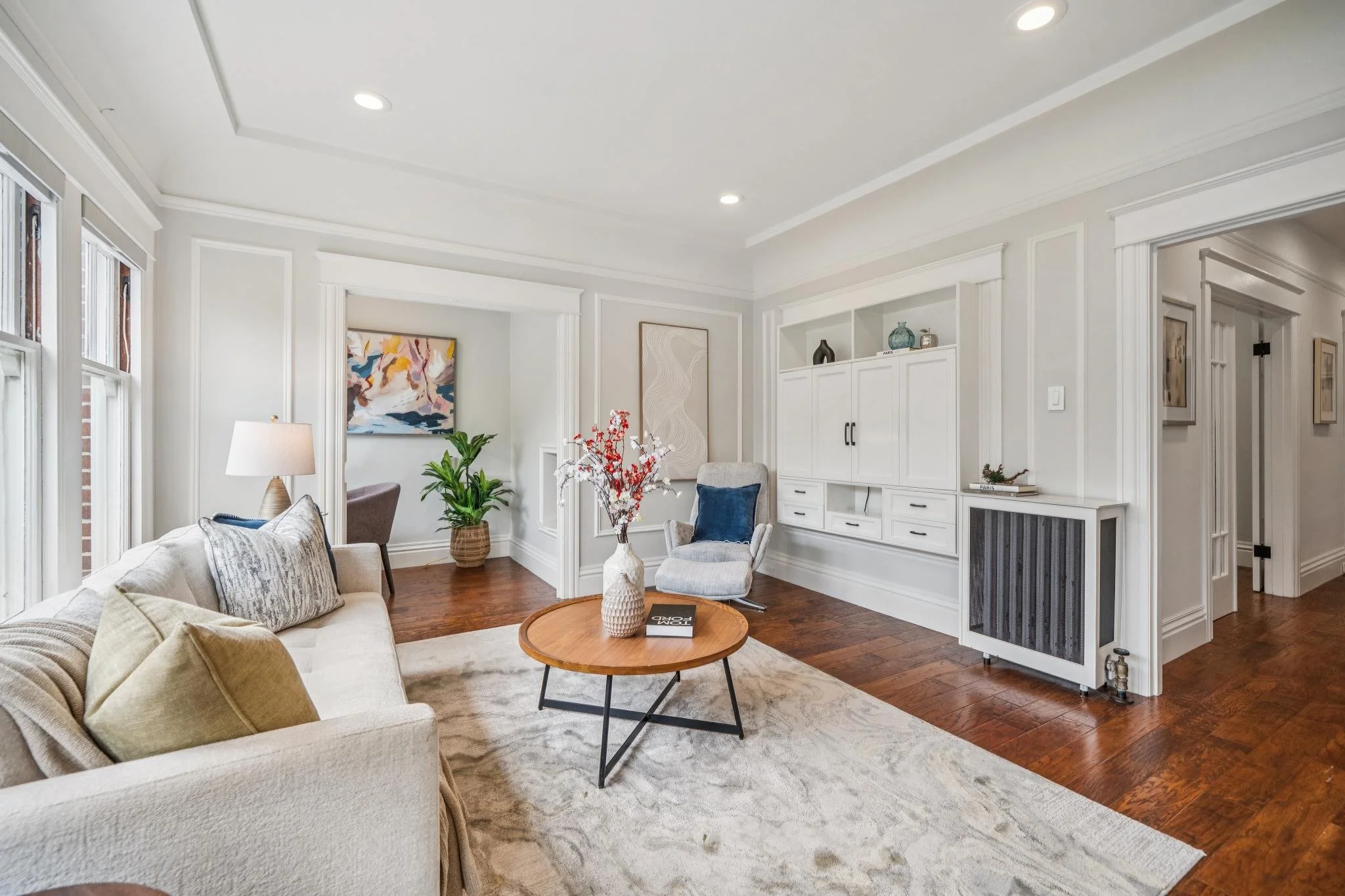 Bright living room with beige sofa, white built-in cabinets, artwork, plants, round wooden coffee table, and hardwood floors.