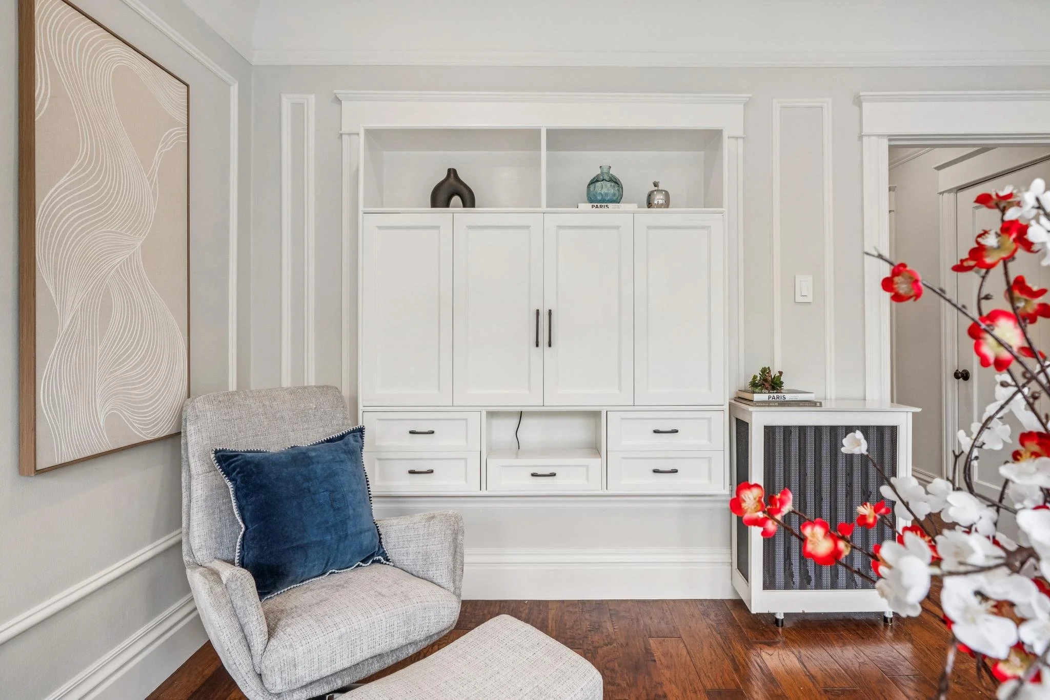 A cozy living room corner with a beige armchair with a blue pillow, a white built-in cabinet with decorative vases on top, a piece of abstract wall art, and a branch with red and white flowers in the foreground.
