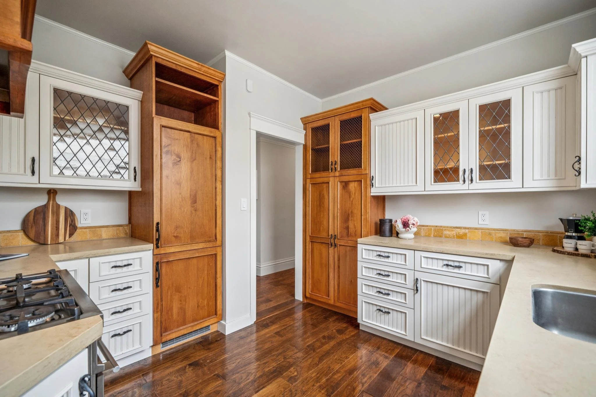 Kitchen with white upper cabinets and natural wood lower cabinets, dark hardwood flooring, beige countertops, and decorative items including a vase with flowers and bowls.