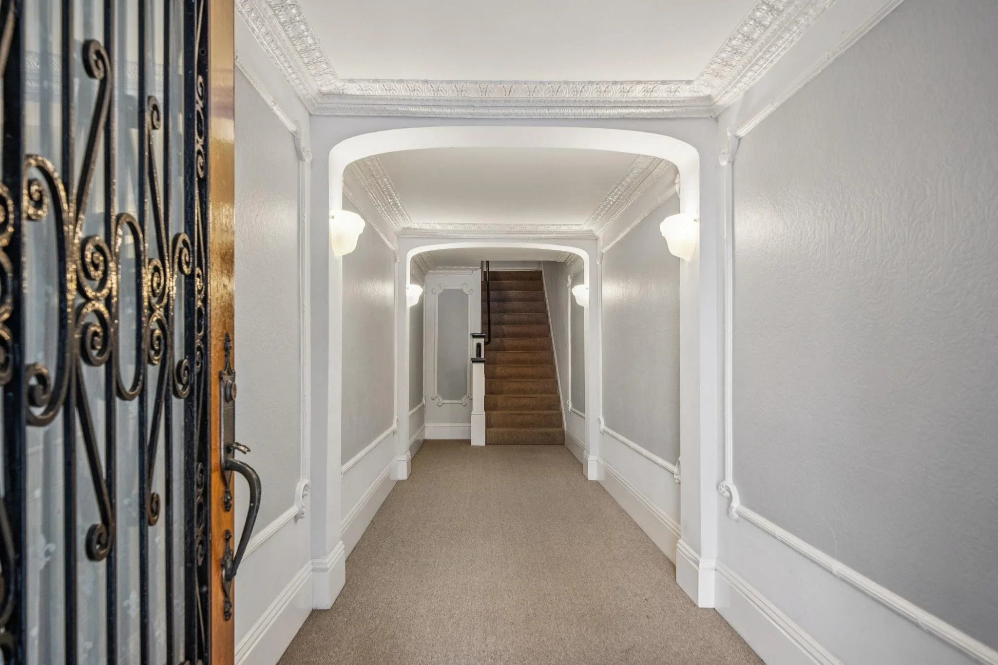 Interior view of a hallway with white walls, ornate crown molding, wall-mounted light fixtures, beige carpet, and a staircase at the end.