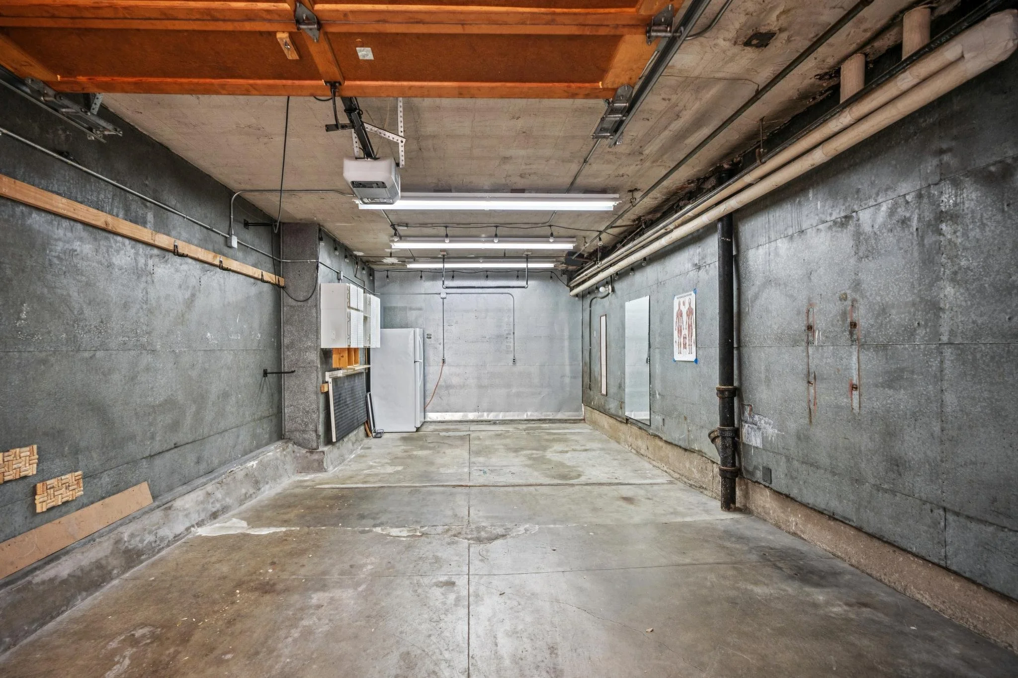Empty garage with concrete walls, floor, and ceiling, fluorescent lighting, and exposed pipes and electrical conduits.