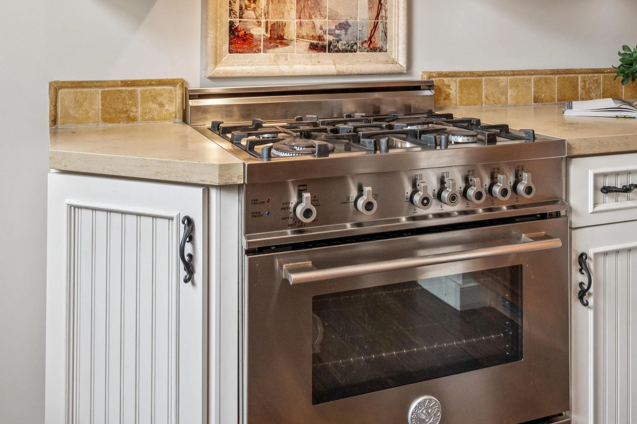 Stainless steel gas stove with six burners in a kitchen with cream and yellow tiled countertops and cabinets, an abstract painting on the wall, and a small potted plant and a stack of papers on the counter.