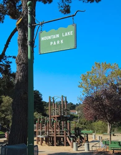 A park sign that reads 'Mountain Lake Park' hanging from a pole, with playground equipment, trees, and a clear blue sky in the background.