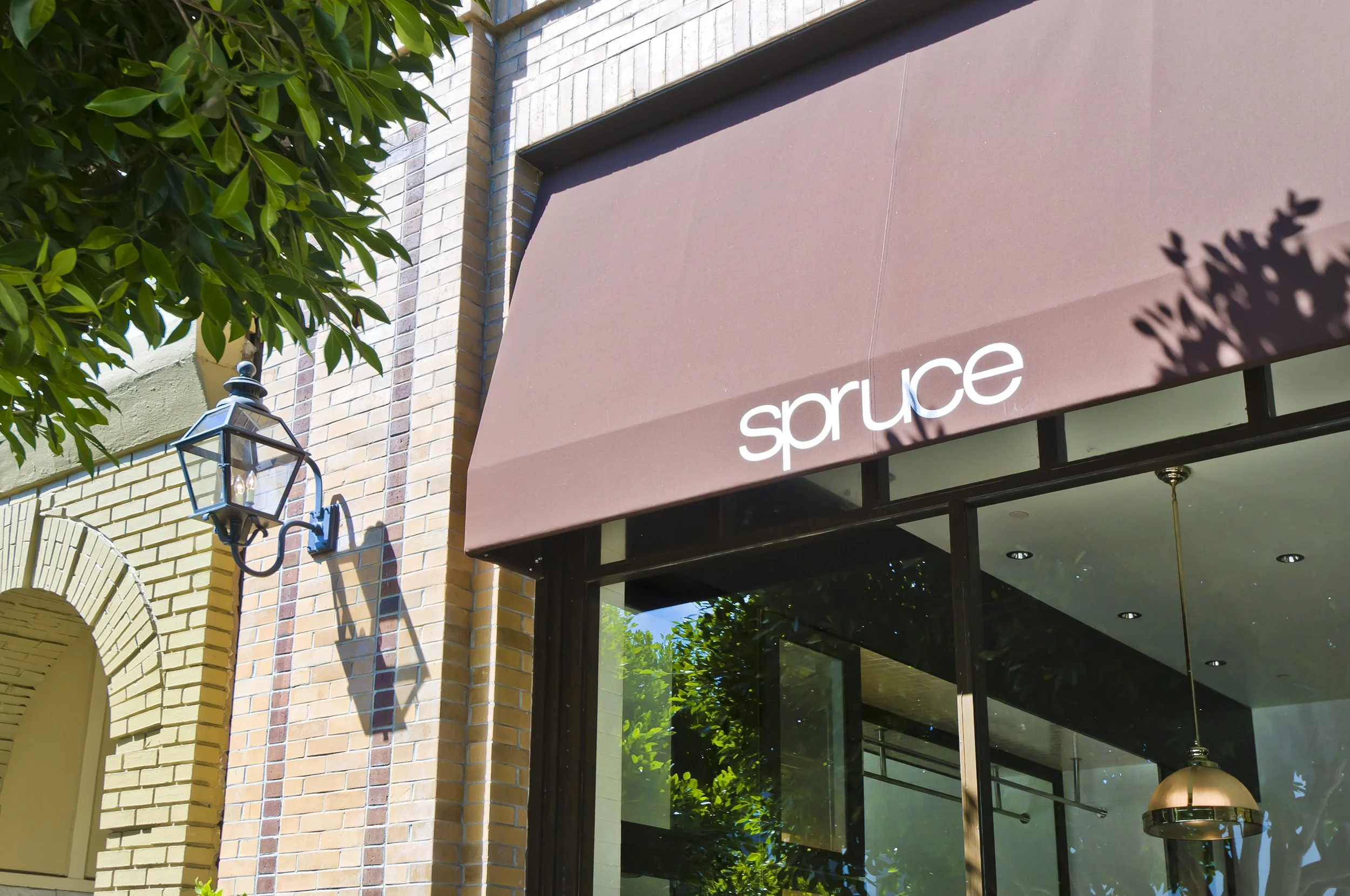 Storefront with a maroon awning that reads 'spruce', a brick building, a black outdoor lantern, and green trees reflected in the large window.