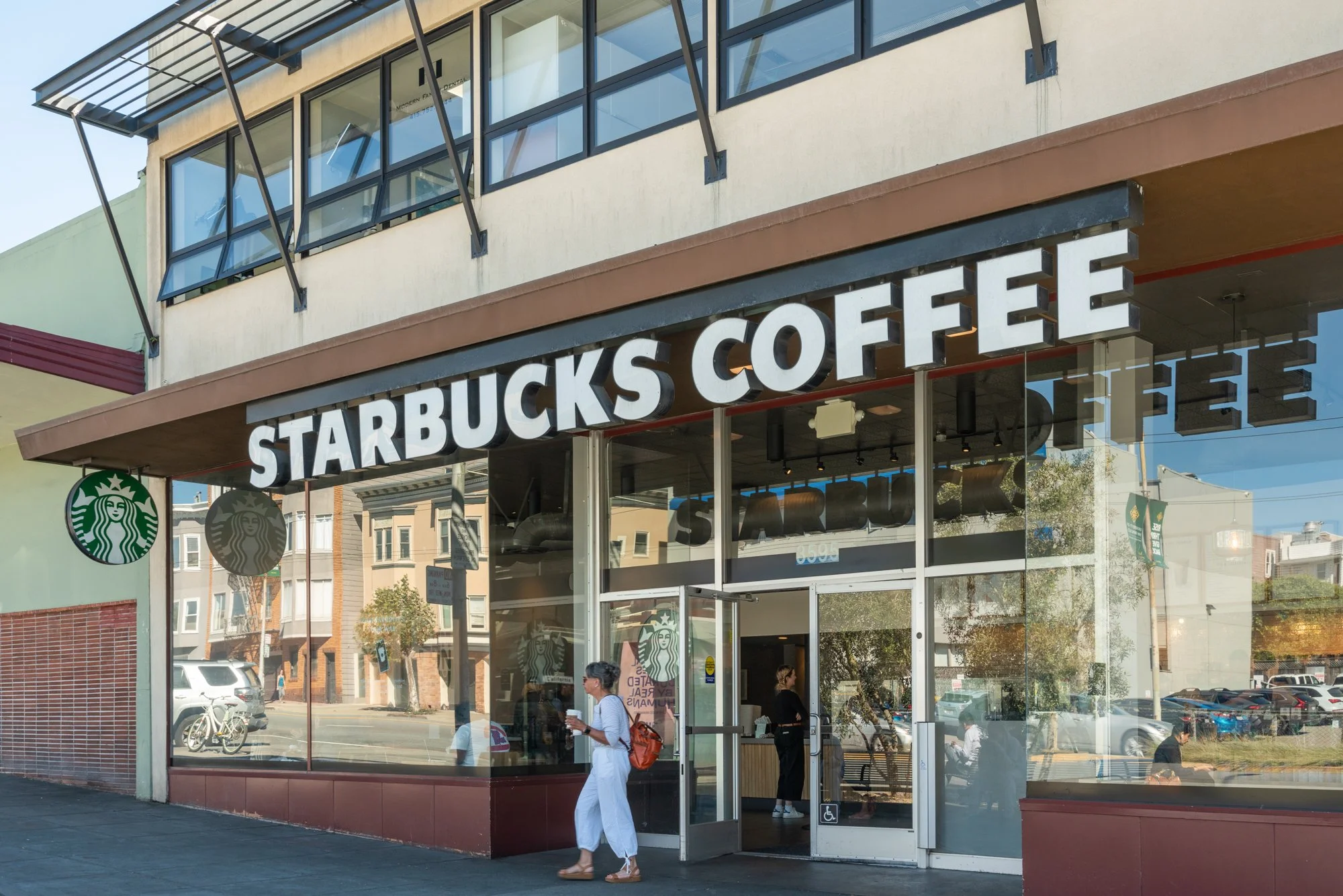 Exterior view of a Starbucks Coffee store with large glass windows and sign, with a person walking in front and a busy street in the background.