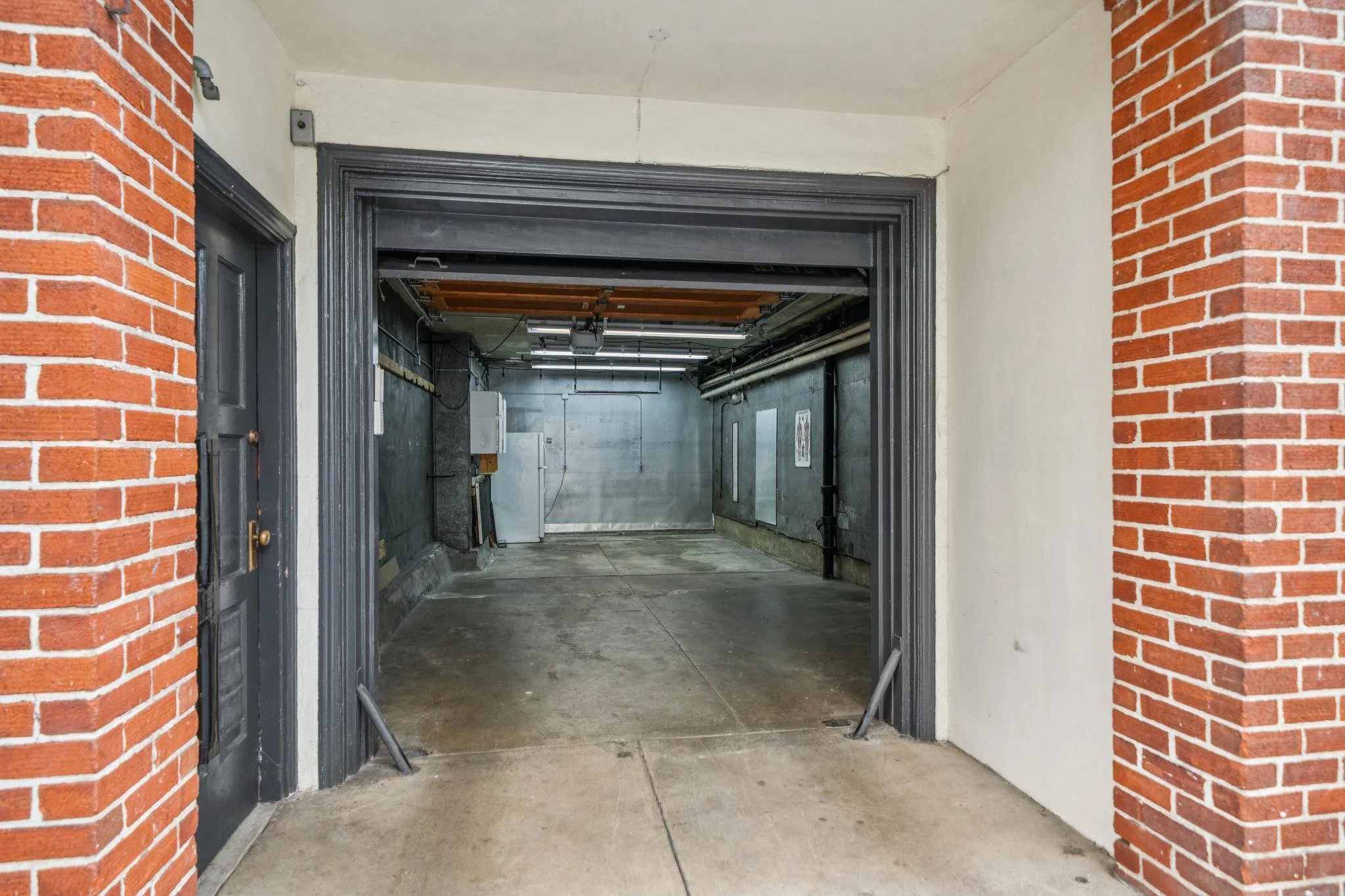 View of an open garage with brick walls on both sides, concrete floor, and partial ceiling visible with exposed pipes and electrical wiring inside.