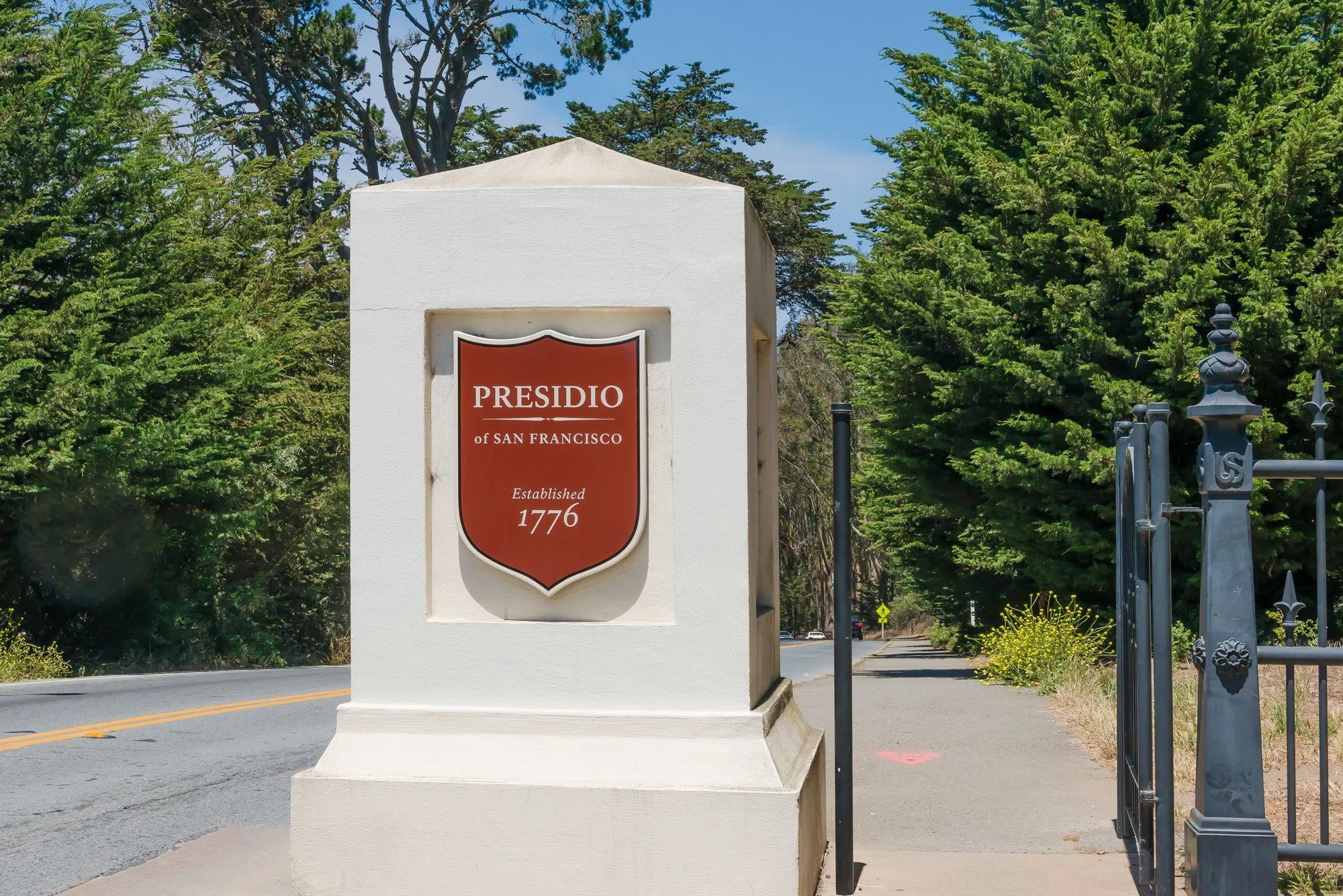 Street view of the entrance to Presidio of San Francisco with a sign on a white concrete pillar, surrounded by green trees, a fence, and a road.