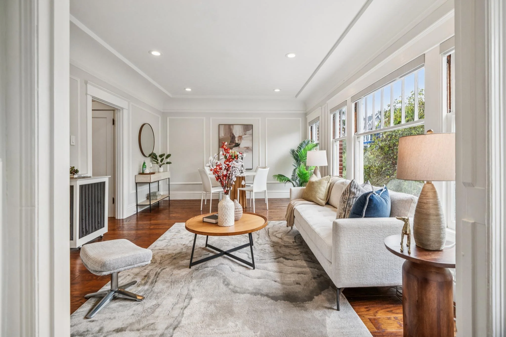 Bright living room with large windows, white walls, wooden flooring, a white sofa with cushions, a round wooden coffee table with vases, a vase with flowers, a side table with a lamp, art on the walls, and indoor plants.
