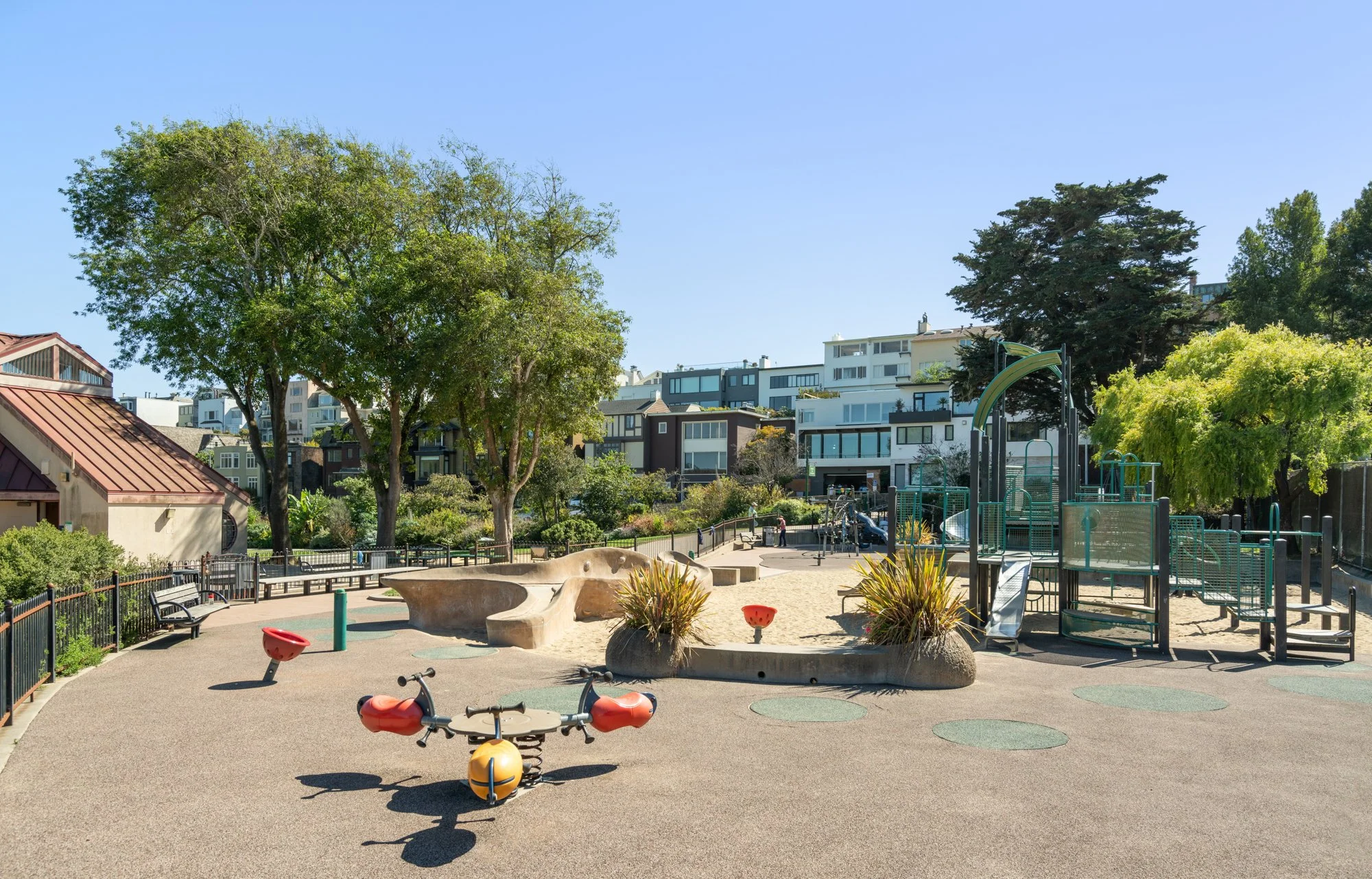 A city park playground with trees, a sandbox, and playground equipment, surrounded by residential buildings under a clear blue sky.