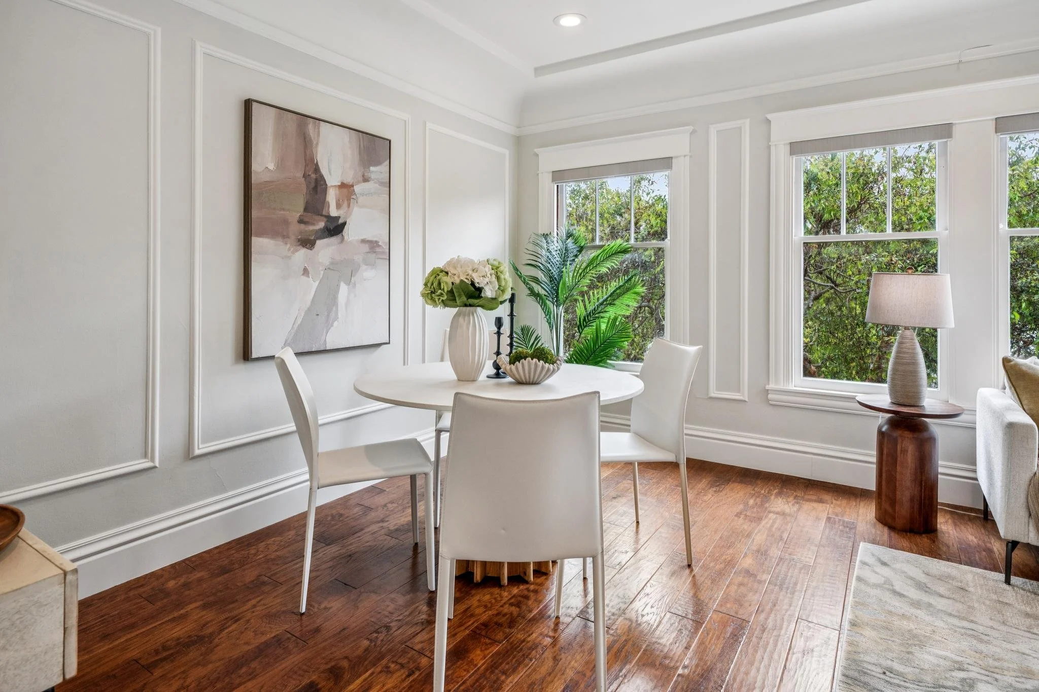 A bright dining room with white walls, a round white dining table, white chairs, a large abstract painting, and a window with greenery outside.