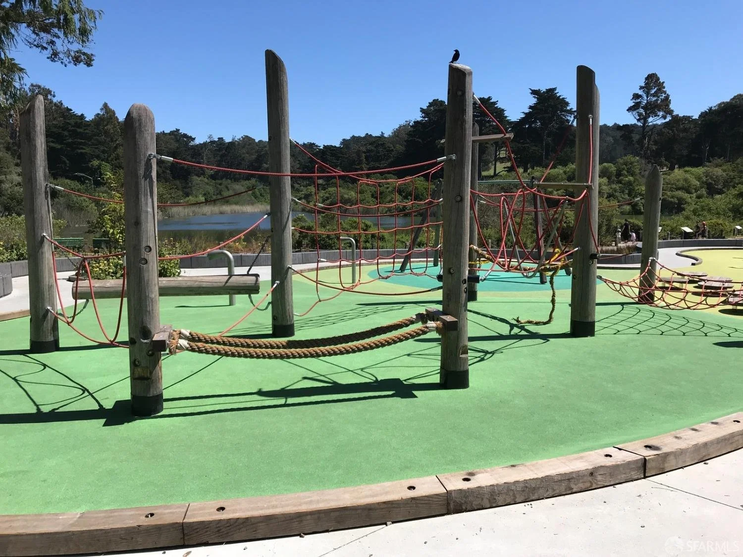 Empty playground with climbing ropes and wooden posts, green rubber flooring, surrounded by trees and a body of water in the background, under a clear blue sky.