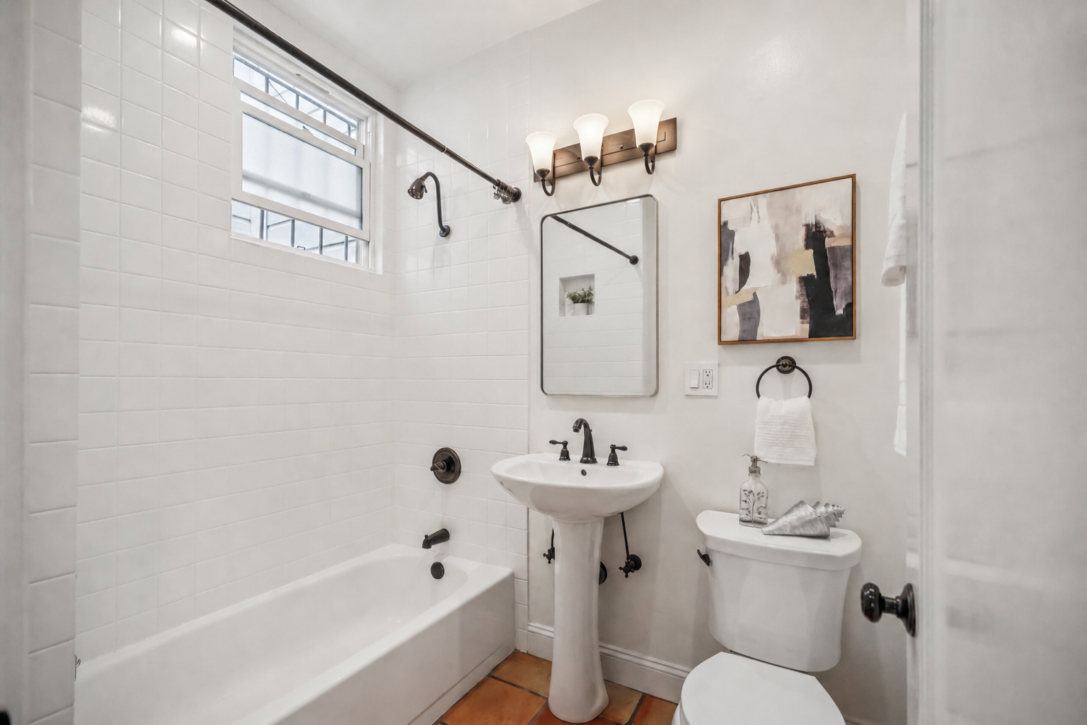 Small bathroom with white tiled walls, a bathtub, a pedestal sink, a toilet, and a window with frosted glass. There is a mirror above the sink, a wall art piece, a towel ring with a white towel, and a decorative bottle on the toilet tank. The lightin
