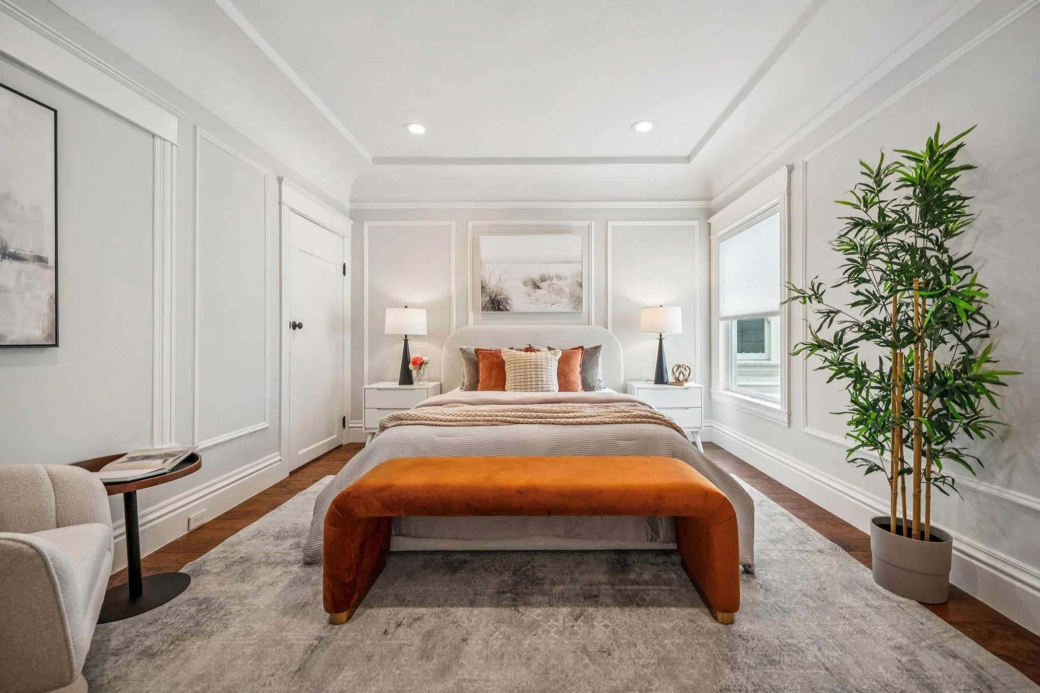 A modern bedroom with white walls, a bed with multiple pillows, two bedside lamps, and a large plant on the right side.