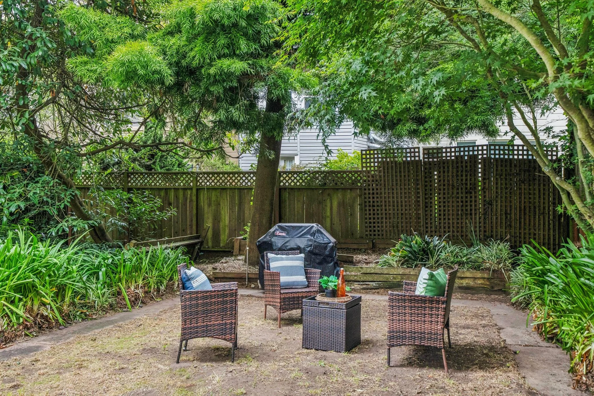 A backyard patio with a table, chairs, and a closed bottle, surrounded by lush greenery and trees, with a wooden fence in the background.
