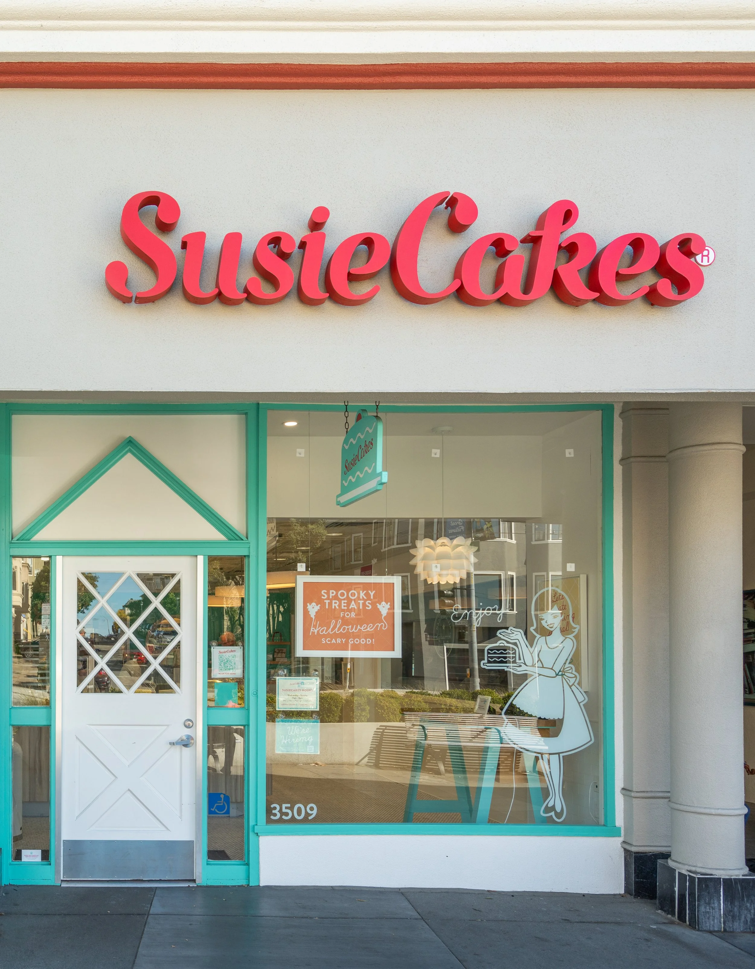 Storefront with a sign that reads 'Susie Cakes' in pink 3D letters. The window has a doodle of a girl holding a tray of cakes, and a sign about Halloween treats.
