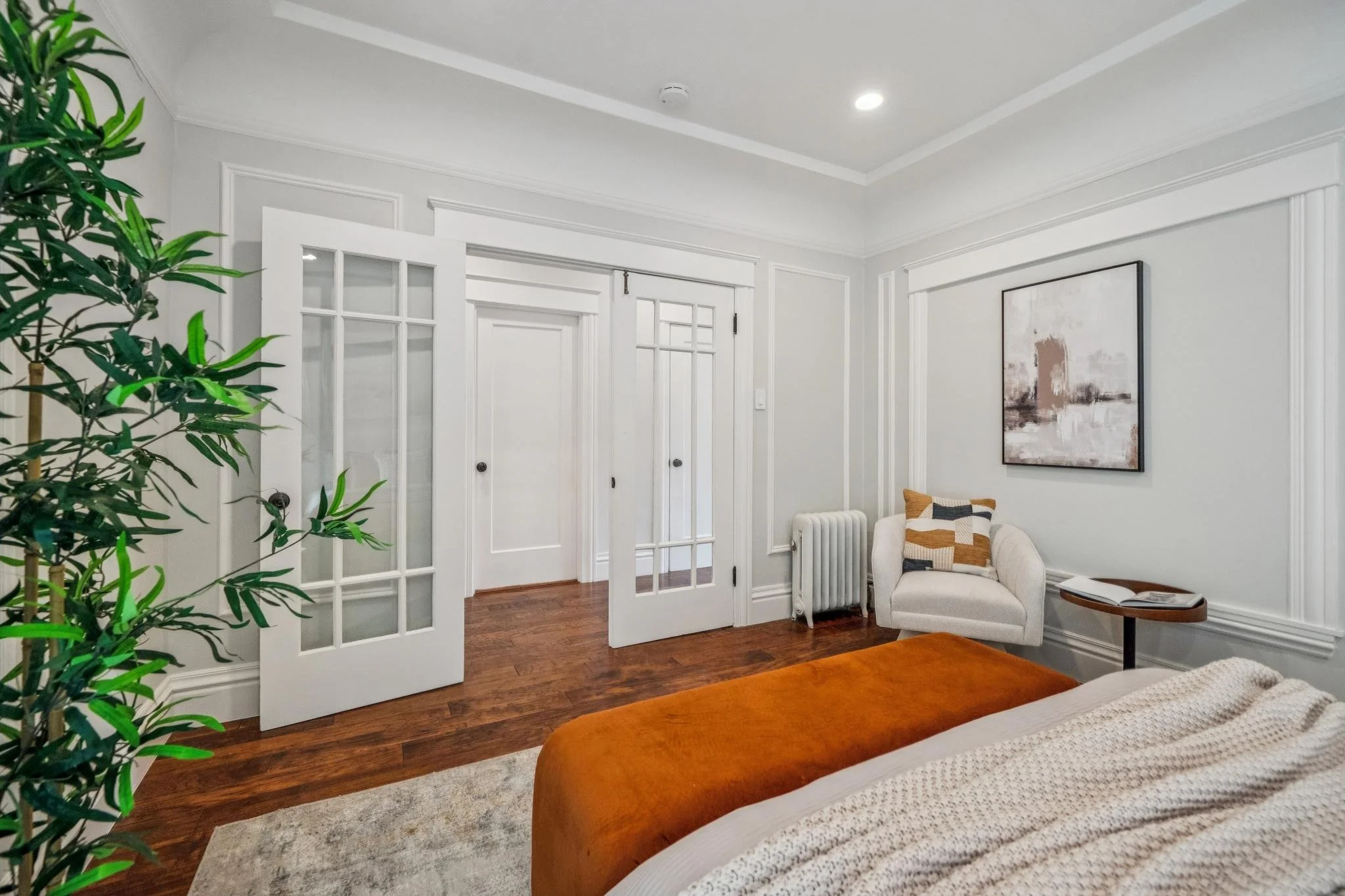 Living room with white walls, hardwood floor, artwork on wall, white armchair with patterned pillow, small round side table with open book, glass-paned doors, potted plant, and ceiling lights.
