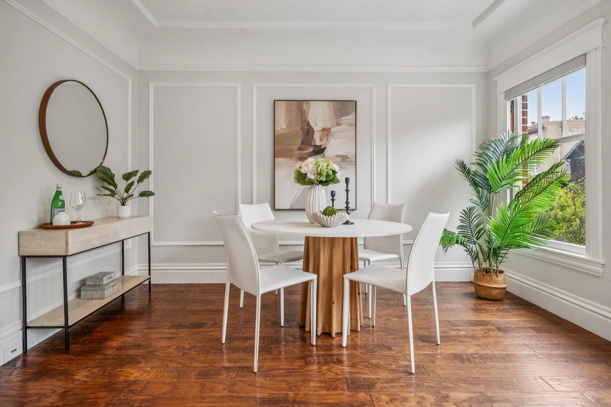 A bright dining room with a round white table, six white chairs, a large potted plant near the window, a sideboard with glassware and a bottle, and a large abstract painting on the wall.