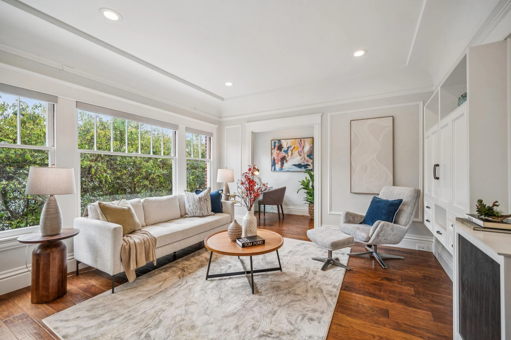 A bright living room with large windows, white walls, and hardwood floors, featuring a beige sofa with pillows, a wooden coffee table with books, a white swivel chair, a side table with a lamp, and abstract artwork on the walls.