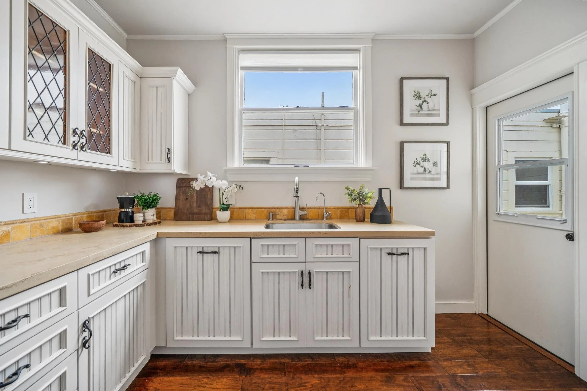 White kitchen cabinets with black handles, a beige countertop, and a window above the sink with a view of a neighboring house and a blue sky, decorated with potted plants and framed botanical prints on the wall.