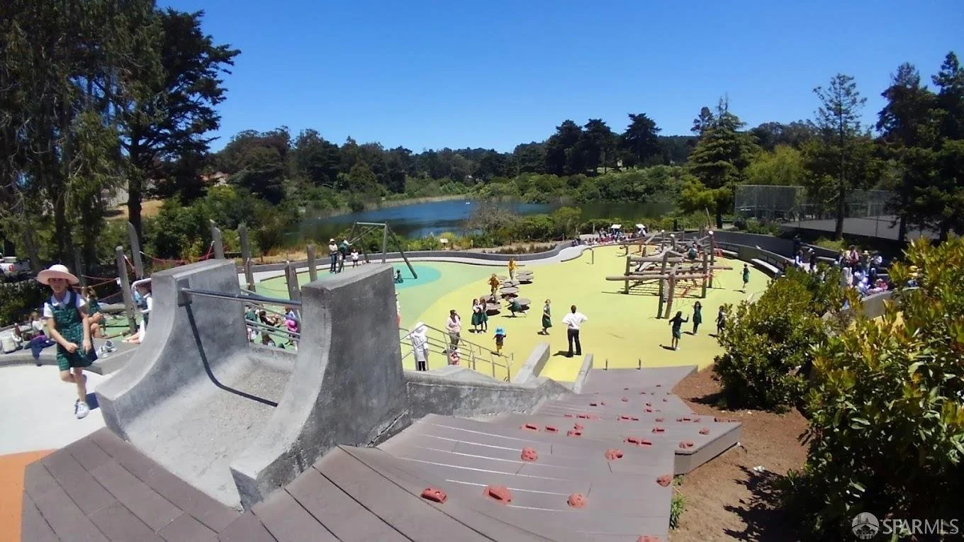 Children and adults playing at a colorful outdoor playground by a lake, with slides, swings, climbing areas, and a rubberized play surface, surrounded by trees and greenery.