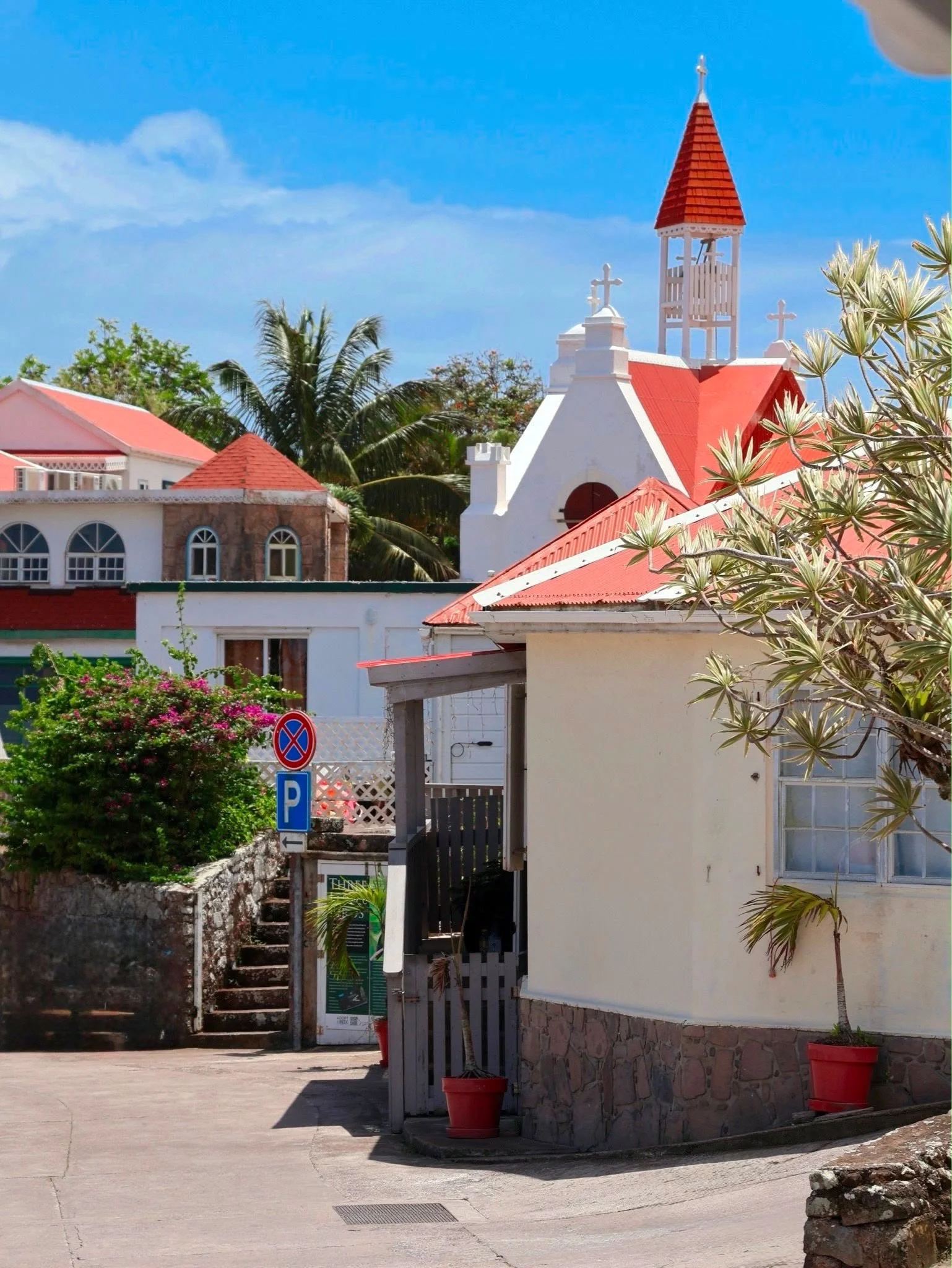 A white church with a red roof and steeple, set against a bright blue sky, surrounded by trees and plants in pots.