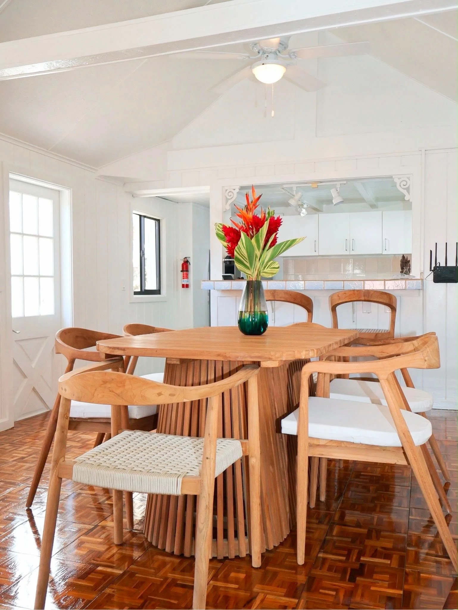 A dining area with a round wooden table, chairs, and a vase with orange and green flowers, in front of a kitchen with white cabinets and a tiled backsplash.