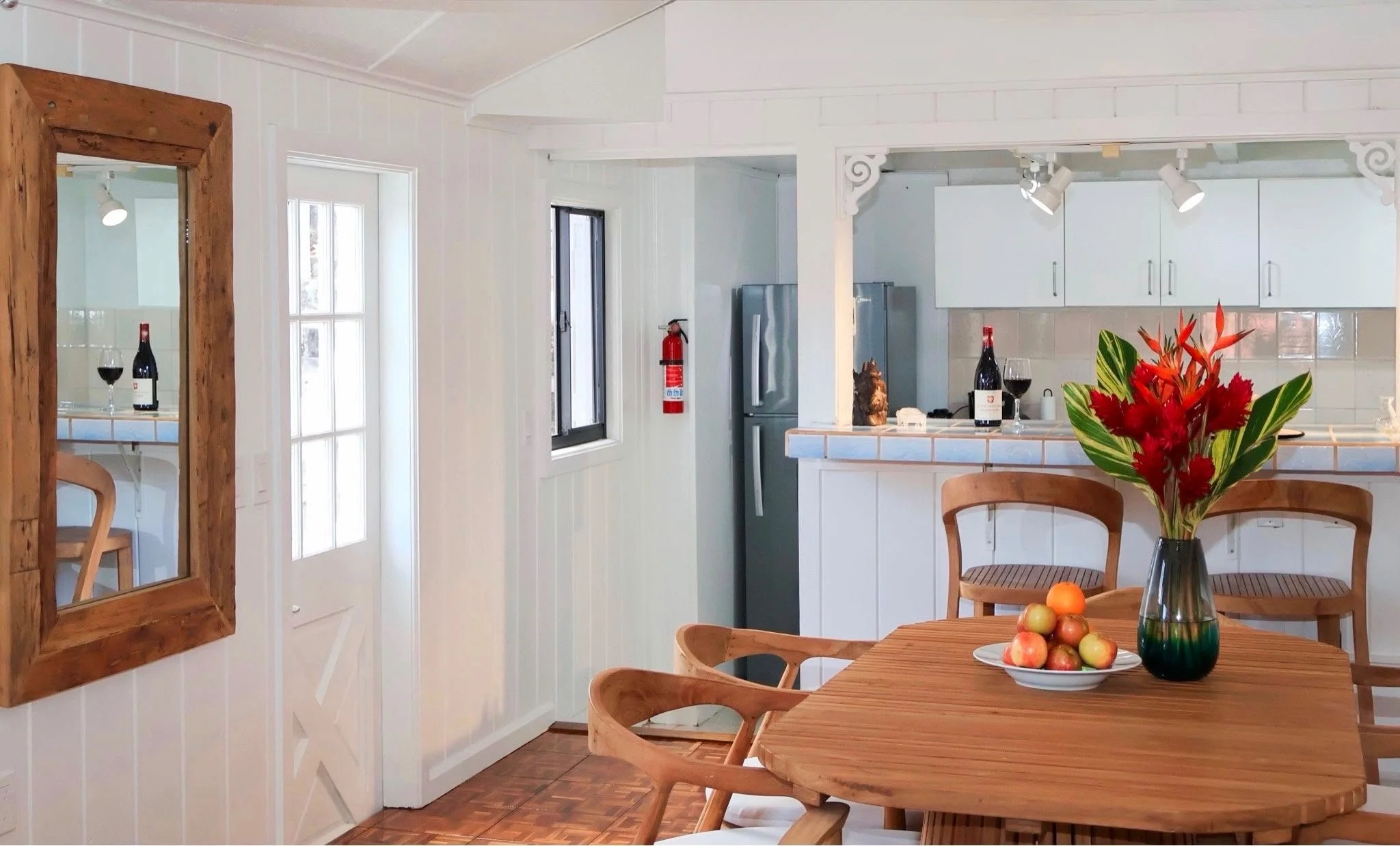 Dining area with wooden table, chairs, vase with red and green flowers, and a bowl of apples. A mirror shows a view of a wine glass and bottle. Kitchen with white cabinets, gray refrigerator, and a fire extinguisher on the wall.