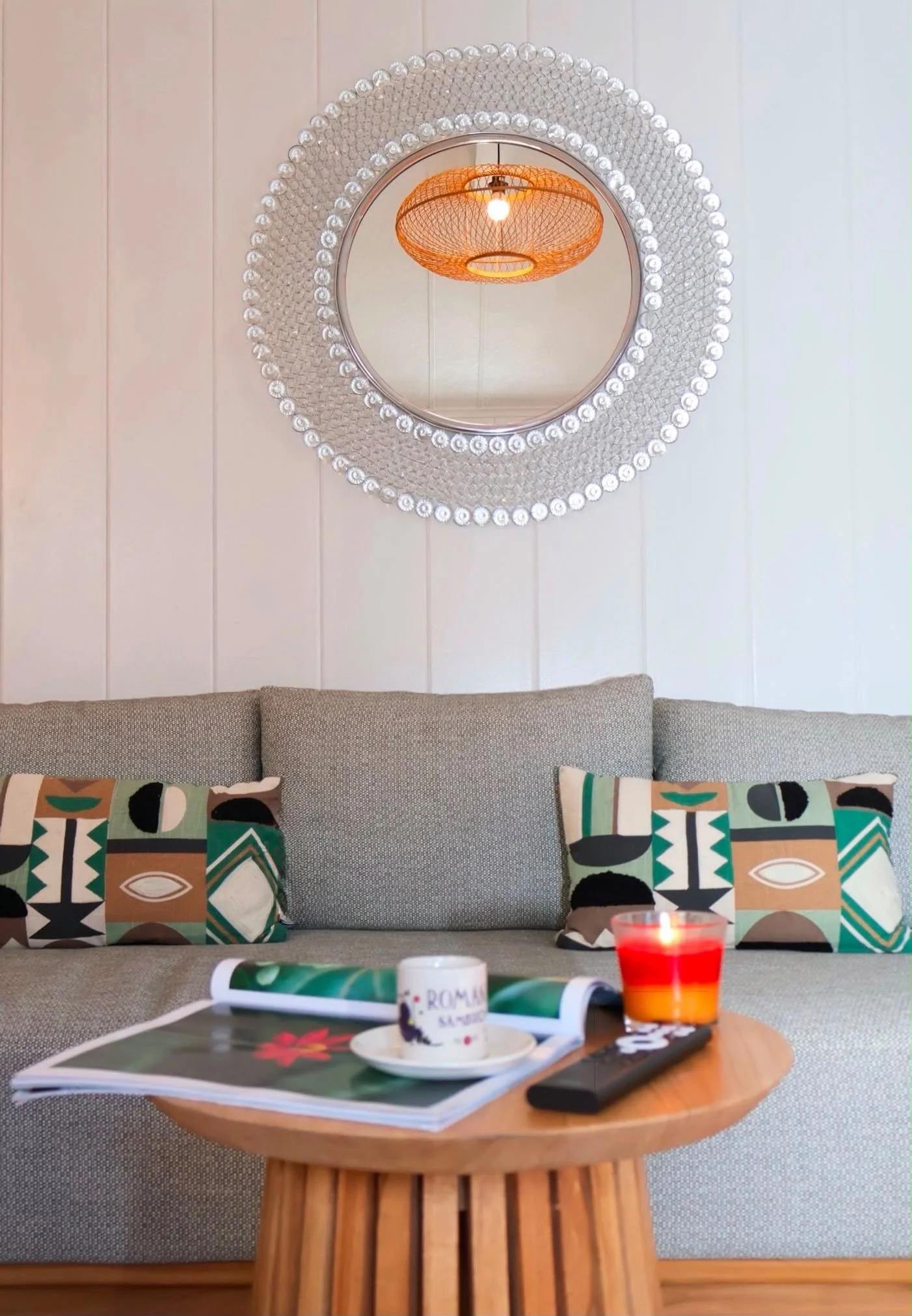 Living room decor including a round mirror with a beaded frame, an orange pendant lamp, a beige sofa with patterned pillows, a wooden table with a magazine, a red candle, and a coffee mug on it.