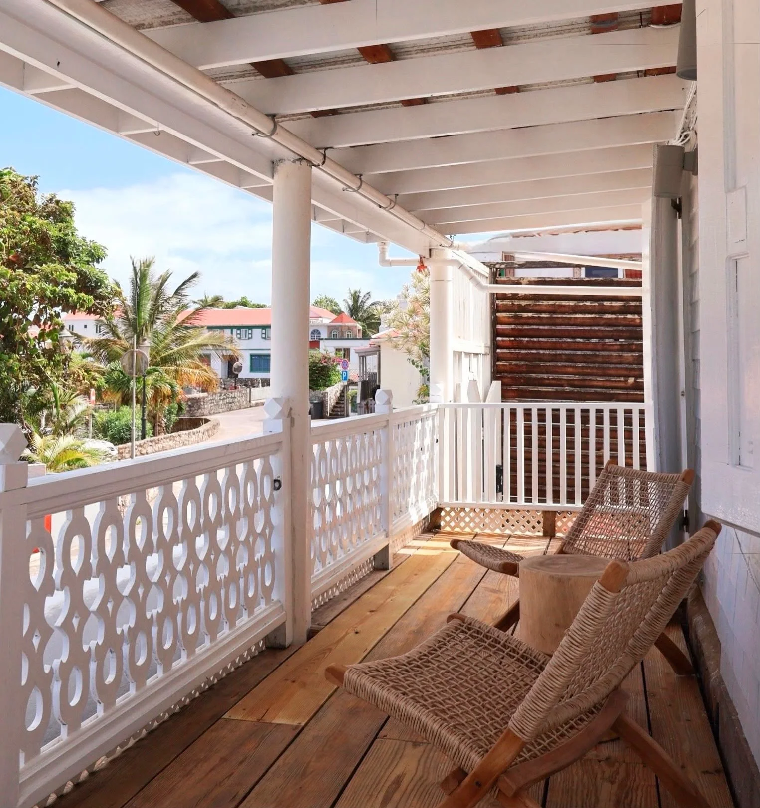 A cozy outdoor balcony with wooden flooring, white decorative railing, two rattan chairs, and lush tropical plants visible in the background. The sky is blue with some clouds.