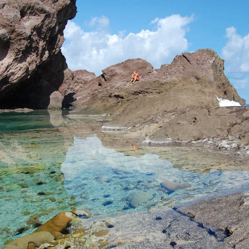 Person sitting on large rocks near clear, shallow water at the shoreline on a cloudy day.