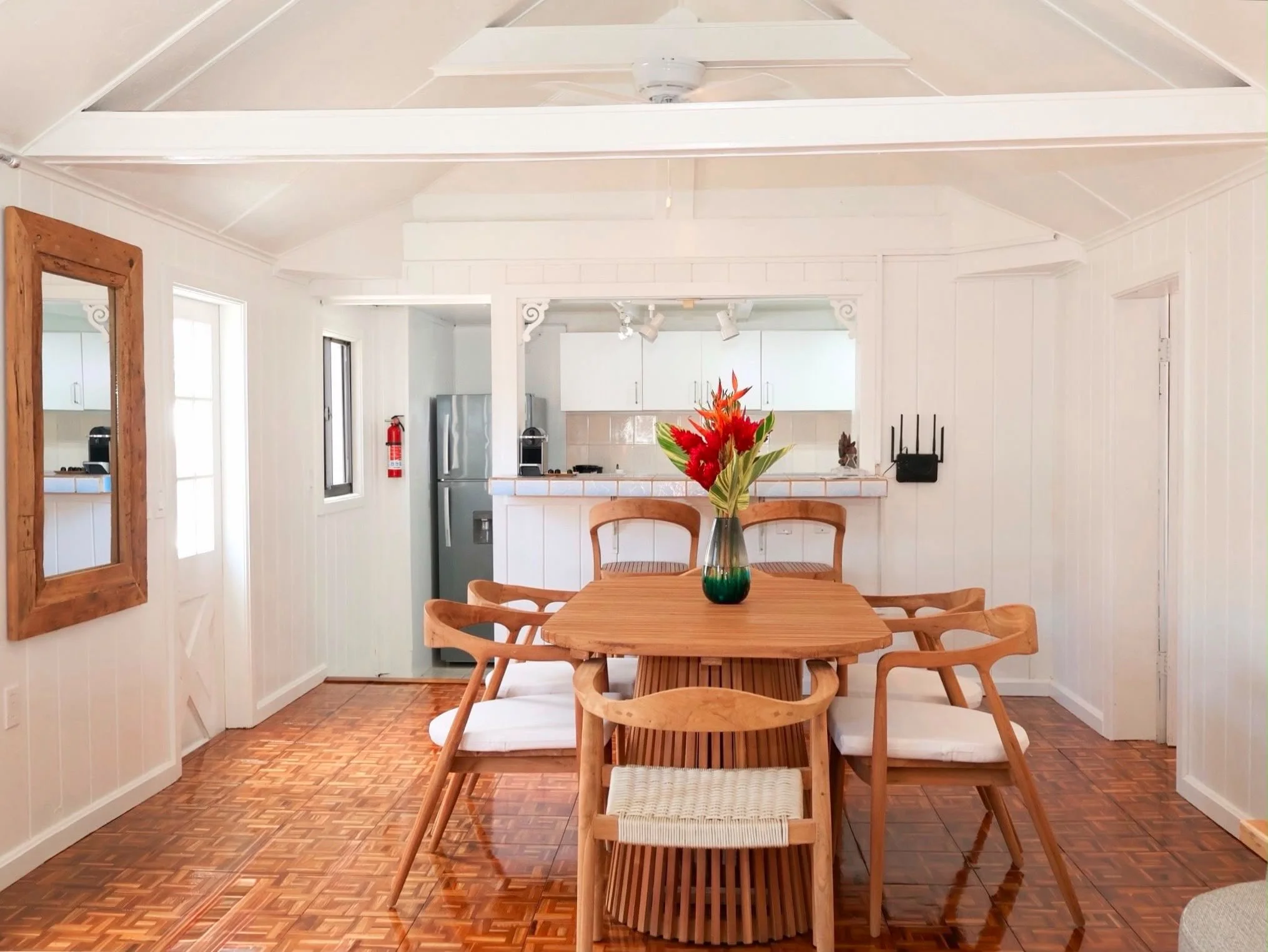 Dining room with a wooden table and chairs, a vase with red and green flowers, white walls, a ceiling fan, and a kitchen area in the background.