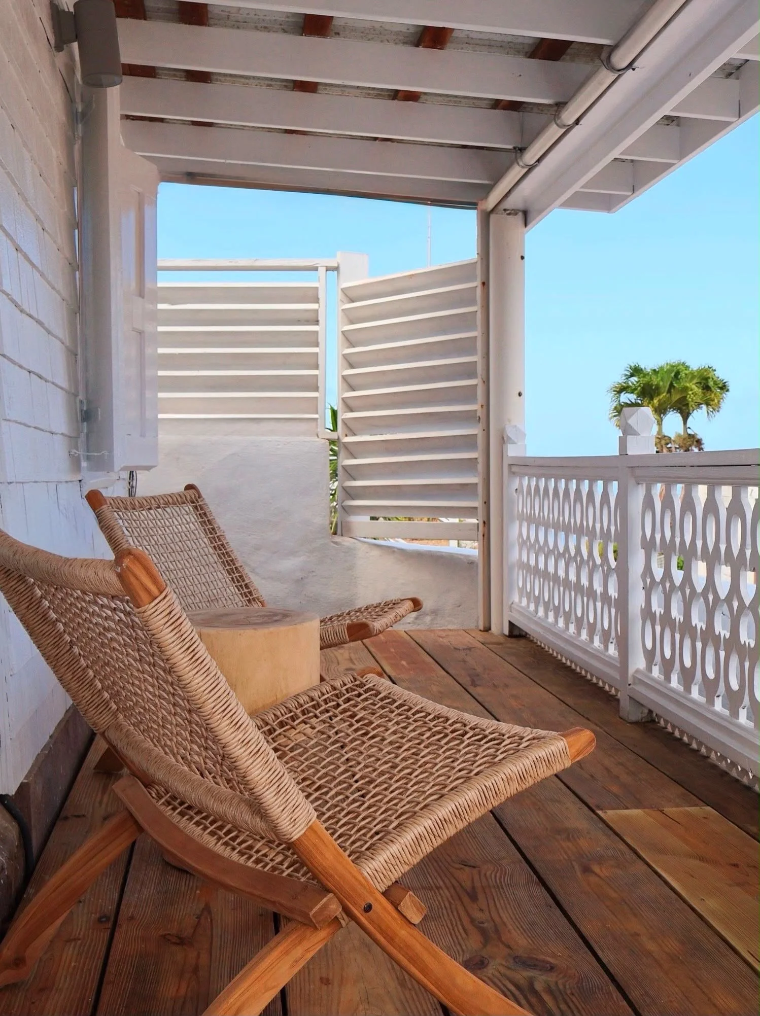 A wooden deck with two wicker lounge chairs and a wooden side table. A white wooden fence with decorative cutouts and a palm tree in the background. Overhead, there is a roof with exposed beams and white pipes.