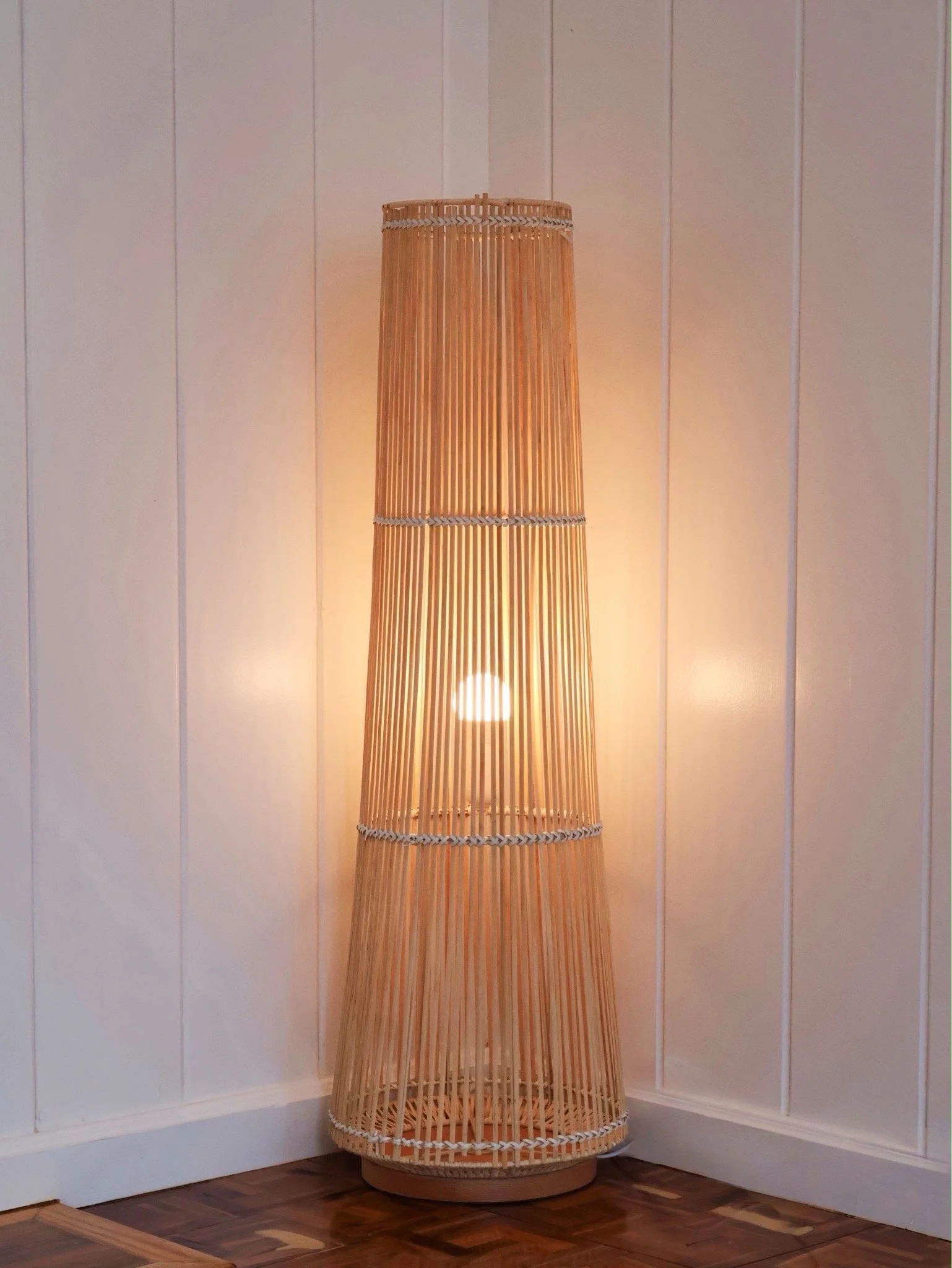 A tall, cylindrical bamboo floor lamp with a woven shade, turned on, against a white paneled wall on a wood floor.
