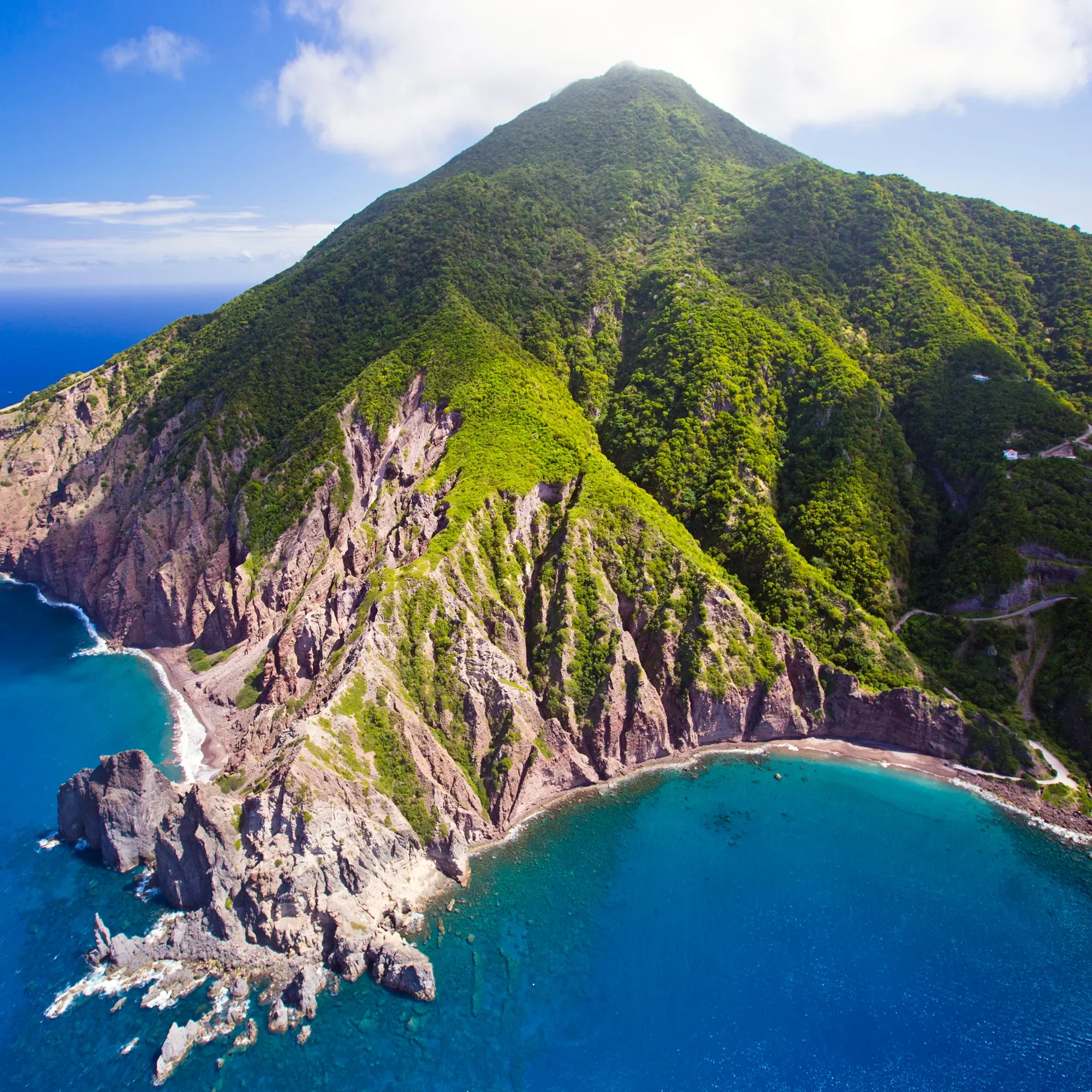 A lush green mountain with a conical shape rises above the ocean, with a coastline featuring rocky cliffs and sandy beach at the base.