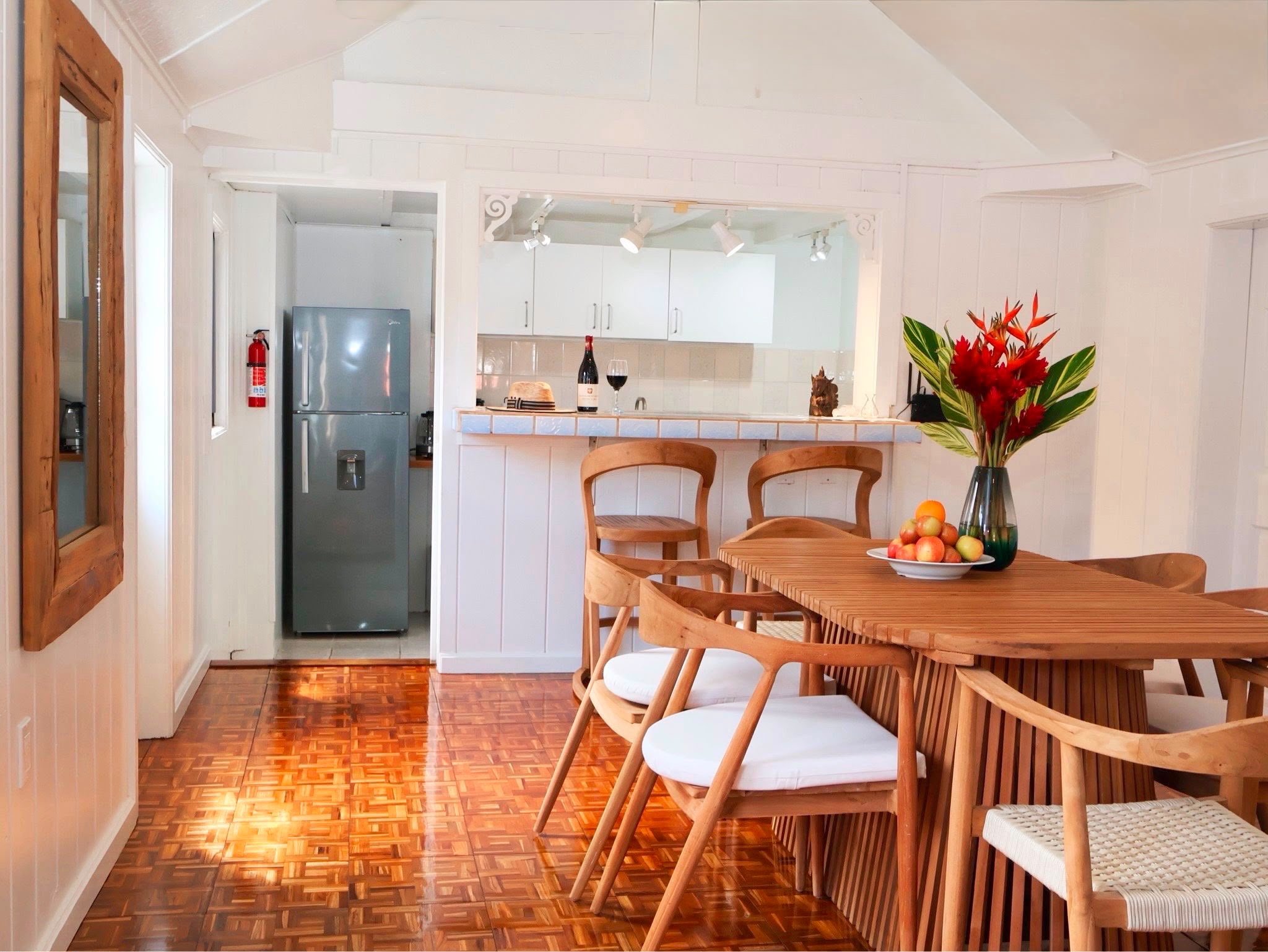 Interior of a kitchen and dining area with wooden furniture, a vase of red flowers, a bowl of apples, a wine bottle, and chairs.