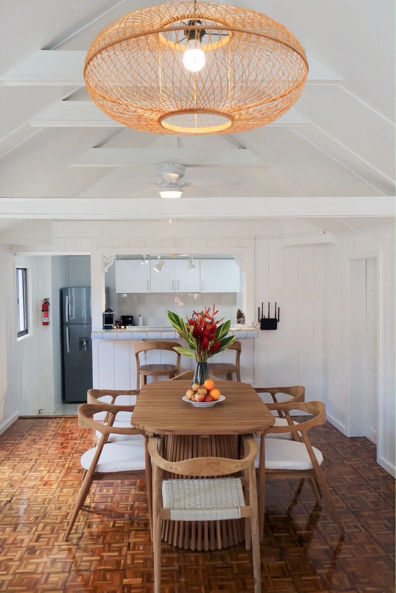 A dining area with a wooden table, six matching wooden chairs with cushions, a vase of tropical flowers and a bowl of apples on the table. Behind, an open kitchen with white cabinets and stainless steel appliances, a hanging light fixture with a woven rattan shade, and a ceiling fan. The floor has a parquet wood design, and the room has white walls and ceiling.